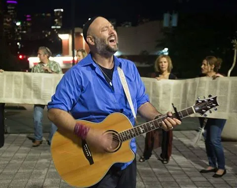 Adult male singer with beard and wearing kippah and a bright blue shirt and black pants, singing with acoustic guitar, and in background is an unrolled torah with adult people holding it.