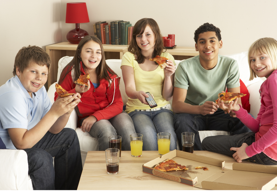 four teenagers of mixed genders and races eating pizza in a living room.