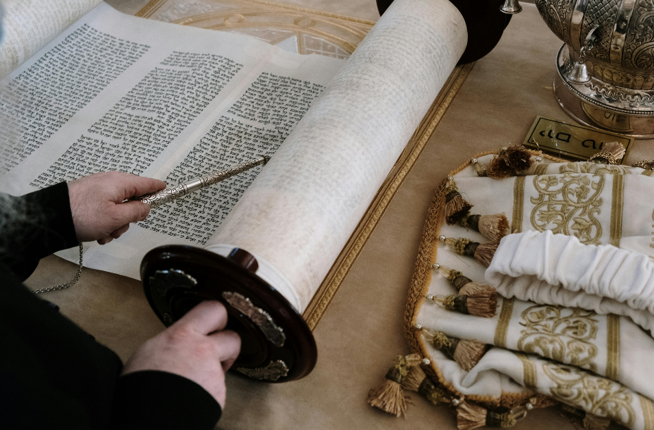 Torah scroll laid out on table, with someone pointing to Hebrew text with a yad with one hand, and holding the wooden scroll handle with the other.  Torah vestments laying to the right of the scroll - silver crown, white and gold cover.
