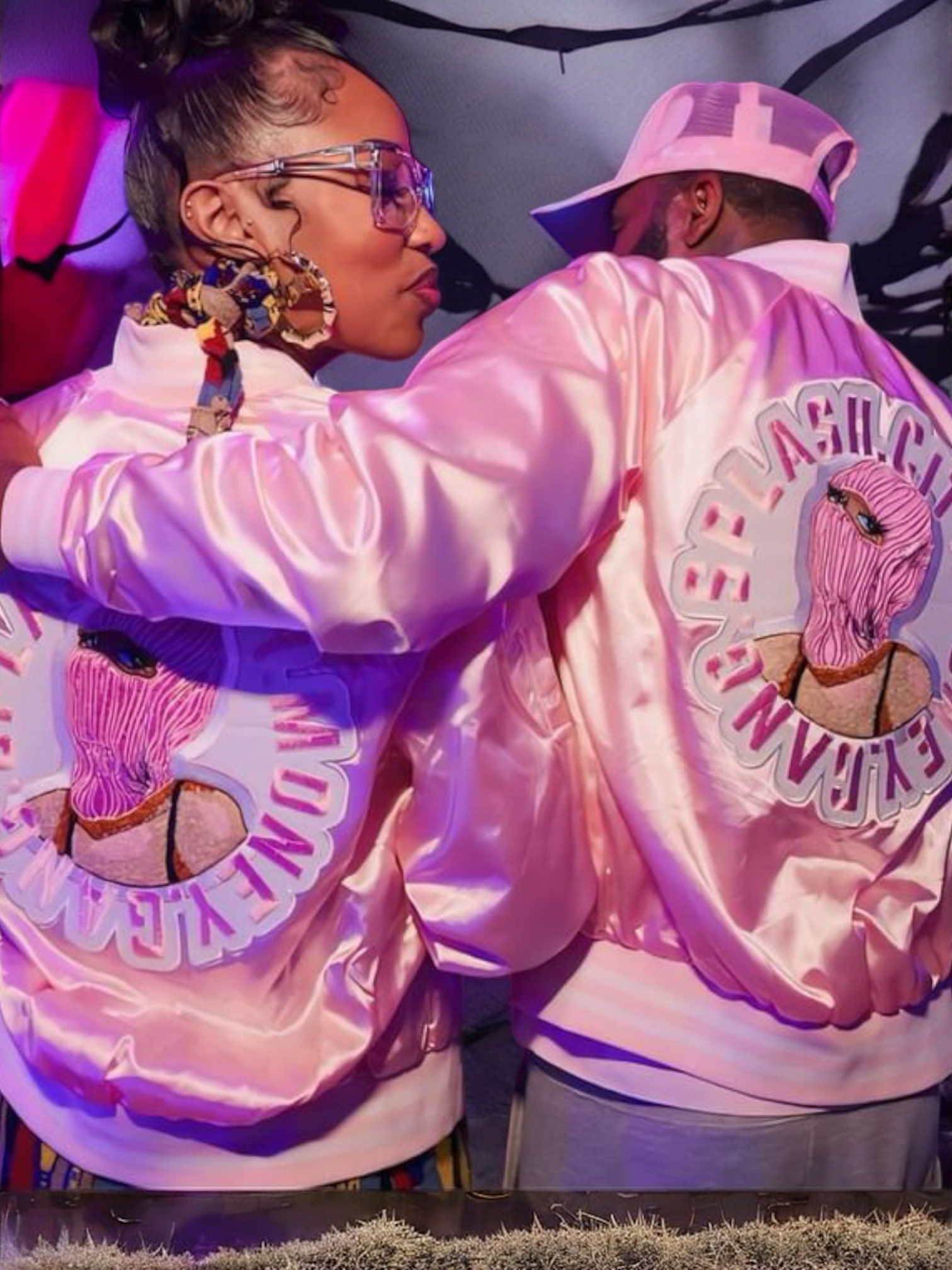 Two people wearing matching pink jackets with a logo featuring a blonde woman with blue eyes and pink hair. One person is wearing glasses and a colorful scarf, and the other has a cap backwards. They are leaning close together.