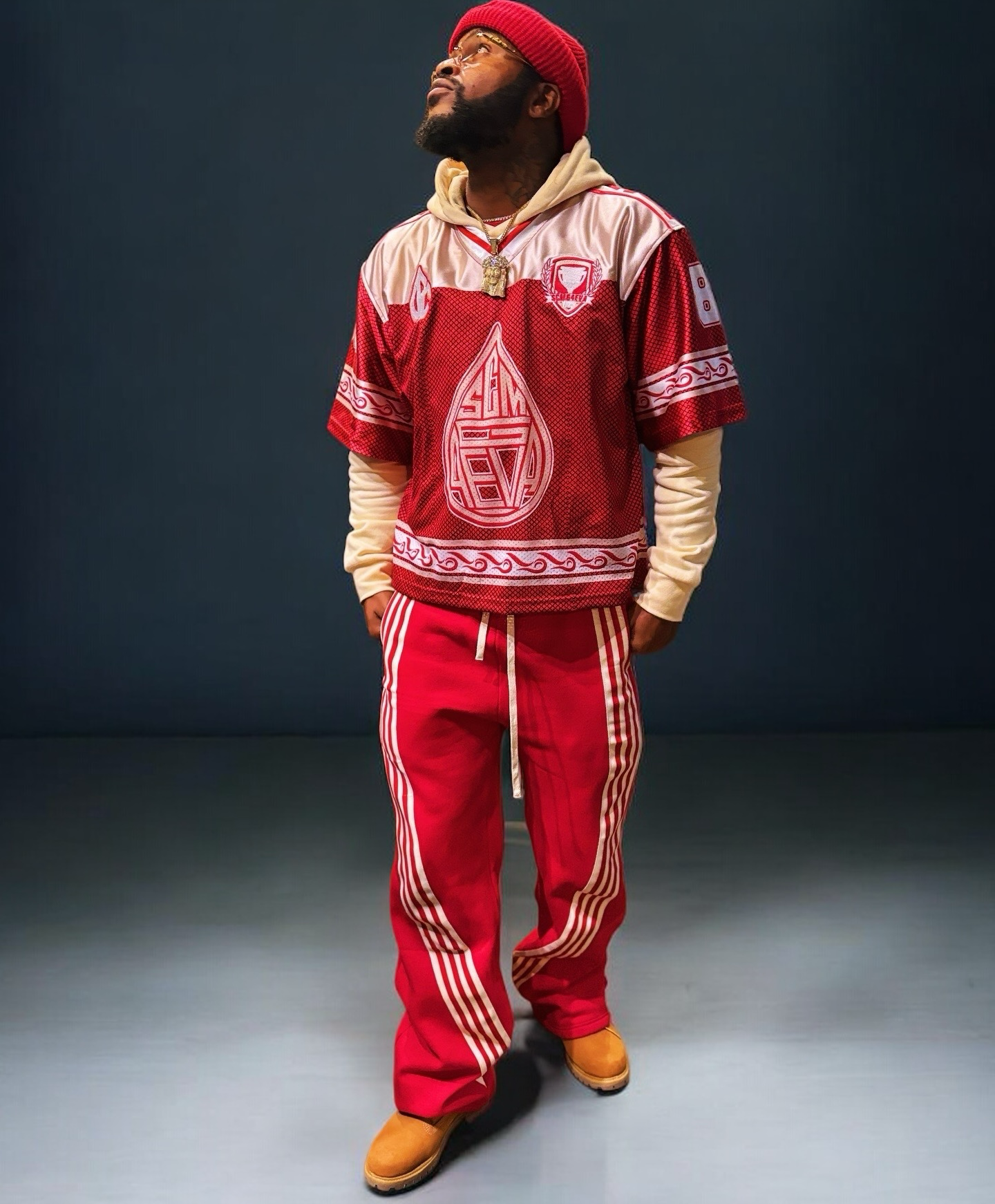 A man with a beard and glasses, wearing a red beanie, beige hoodie, SCMG4EVA SplashSeason Jersey, red pants SCMG4eva WaveRunner pants with white stripes, tan boots, and jewelry, standing against a dark background.