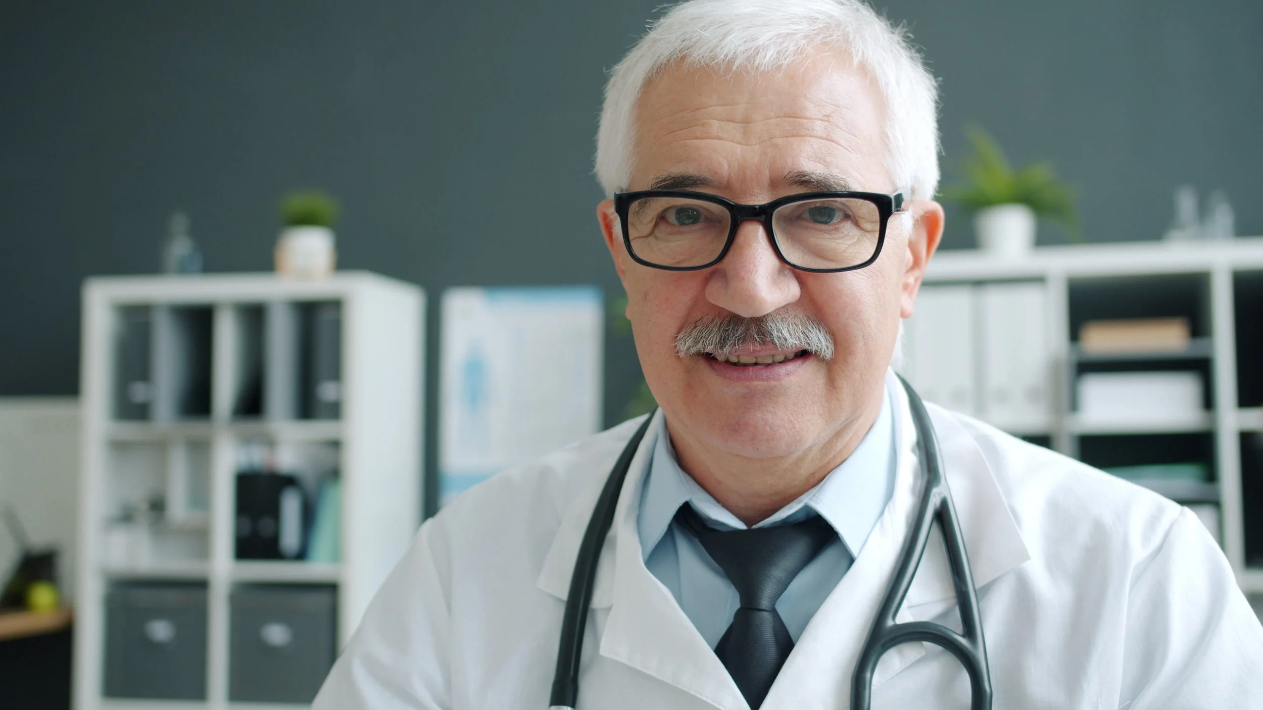 An older doctor with white hair in a white coat standing in an office.