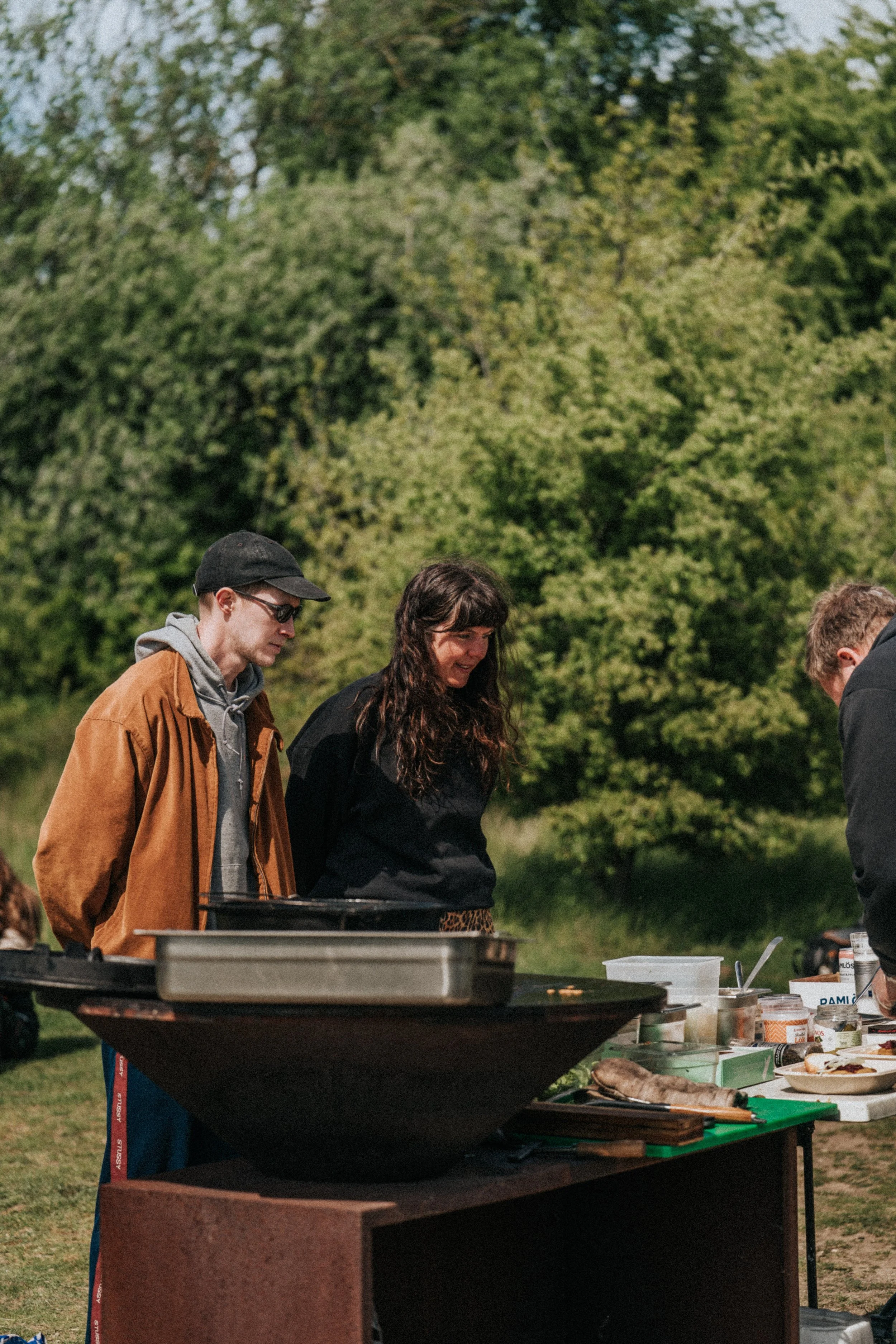 Tre personer står utomhus vid en matlagningsstation med grill och mat. Bakgrund av gröna träd.