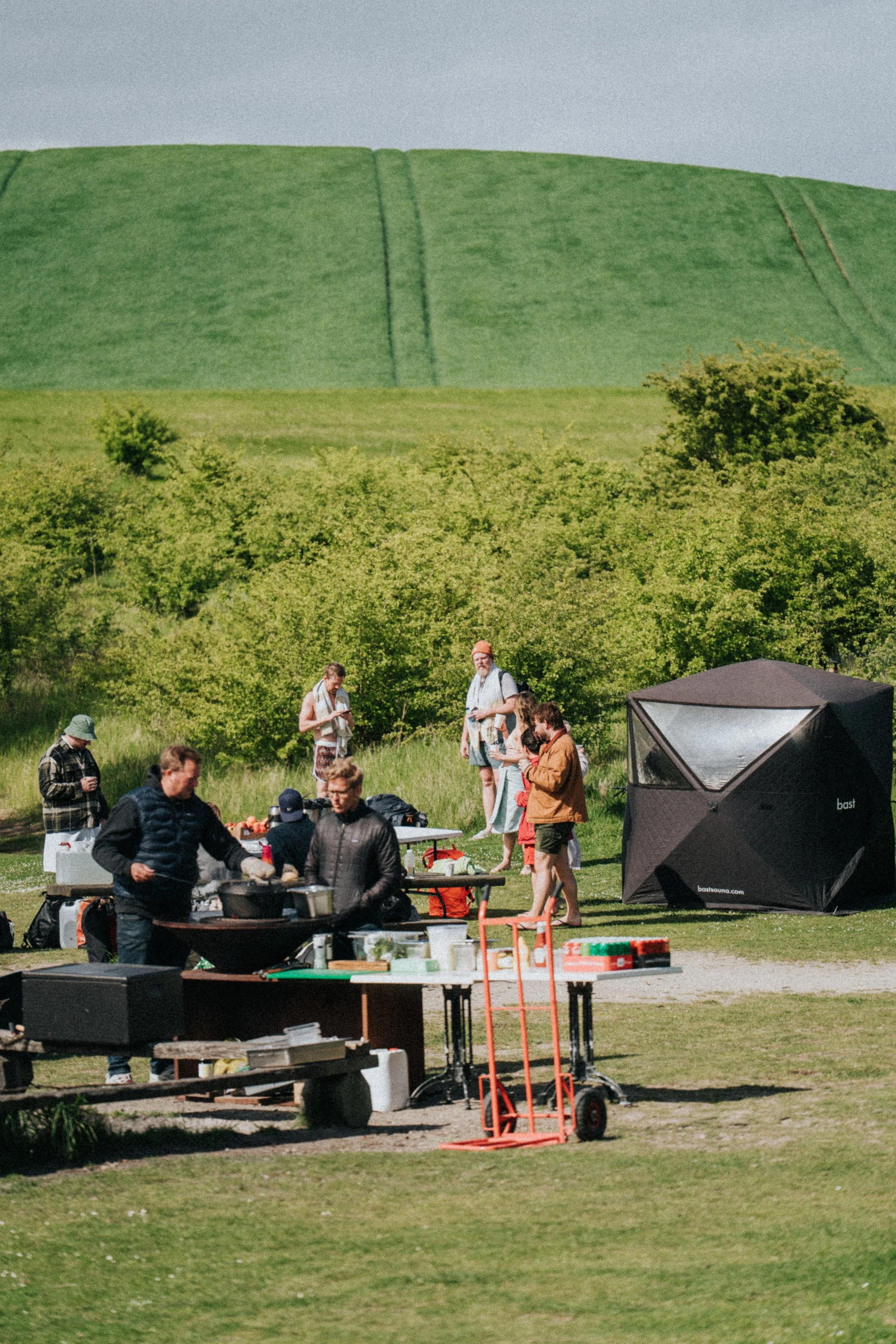 En grupp människor som grillar och umgås utomhus på en gräsplätt med gröna träd och gröna kullar i bakgrunden.