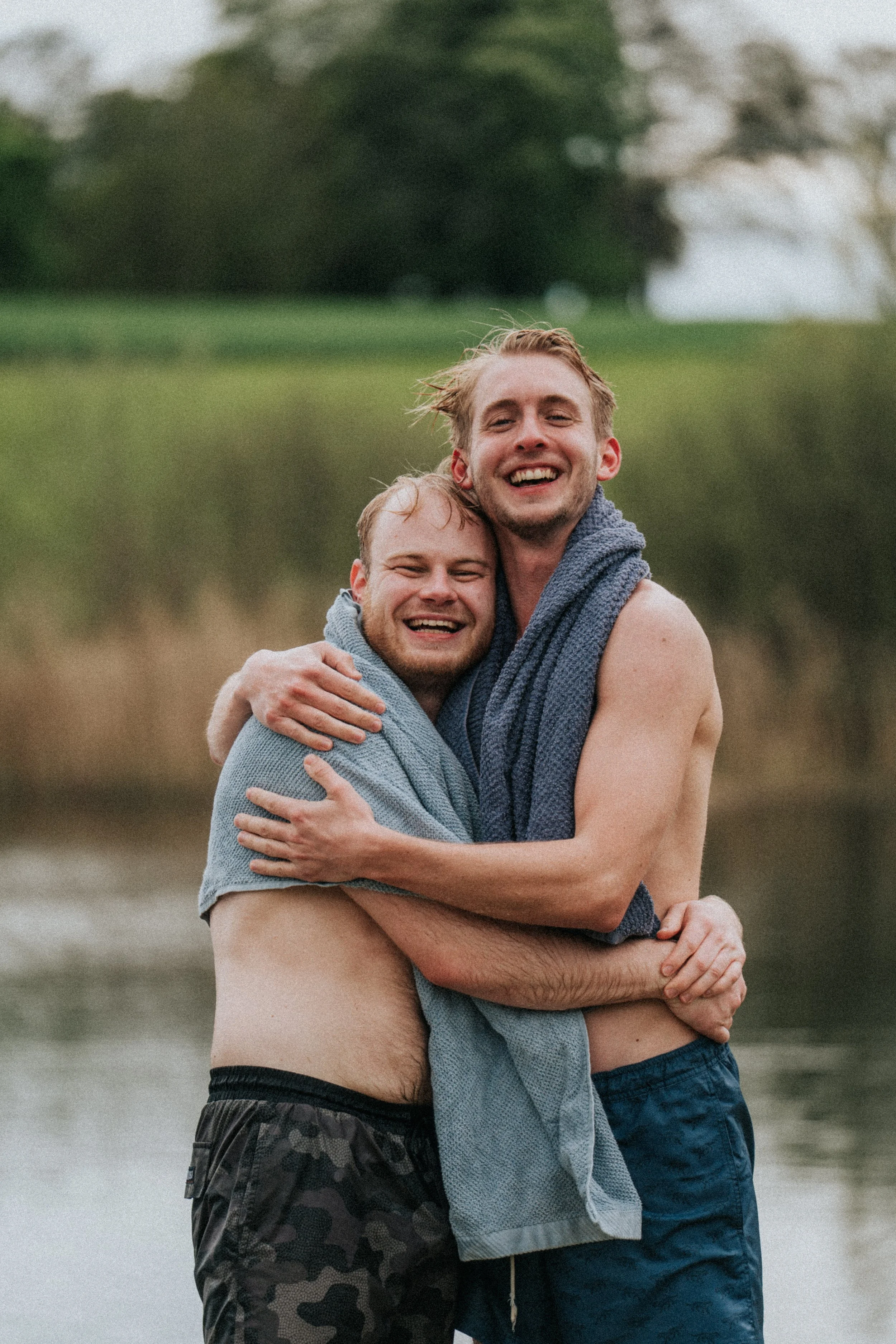 Två män som skrattar och kramas ute vid en sjö med gröna träd i bakgrunden, båda svettiga och utan magtröjor med badrockar.