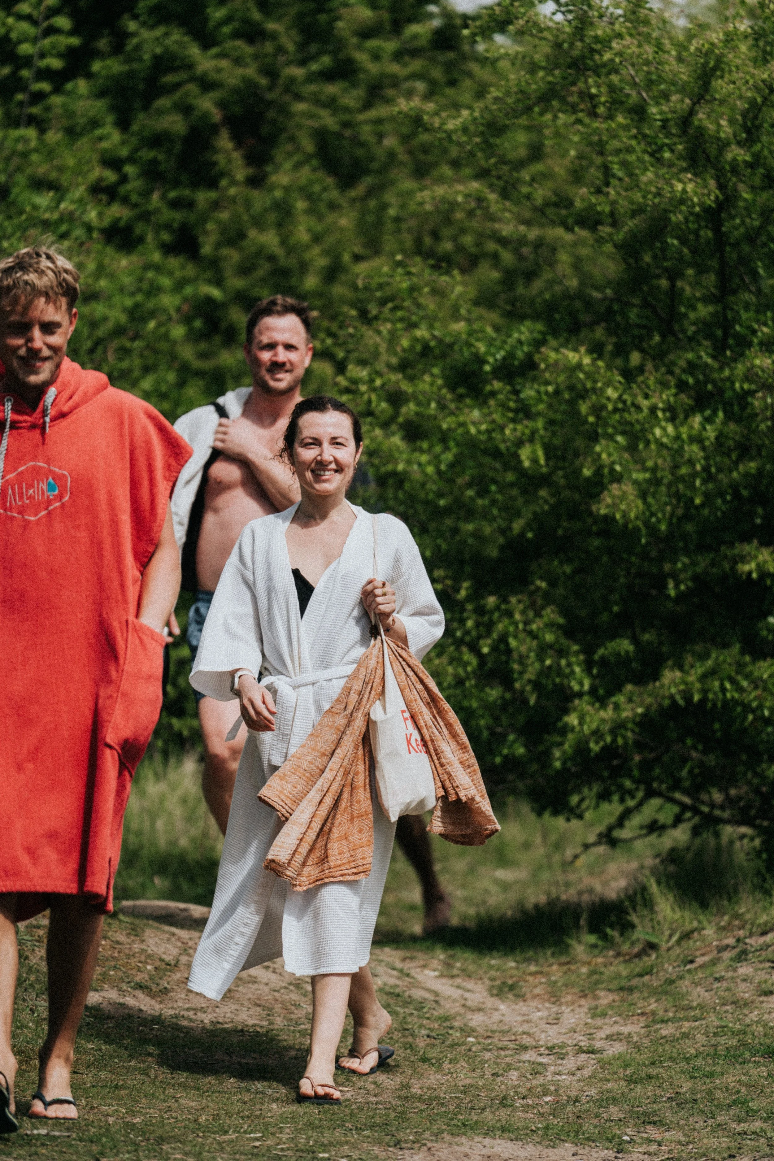 Tre personer promenerar i naturen, en kvinna bär en handduk och en liten väska, de andra två männen bär handdukar och badkläder.