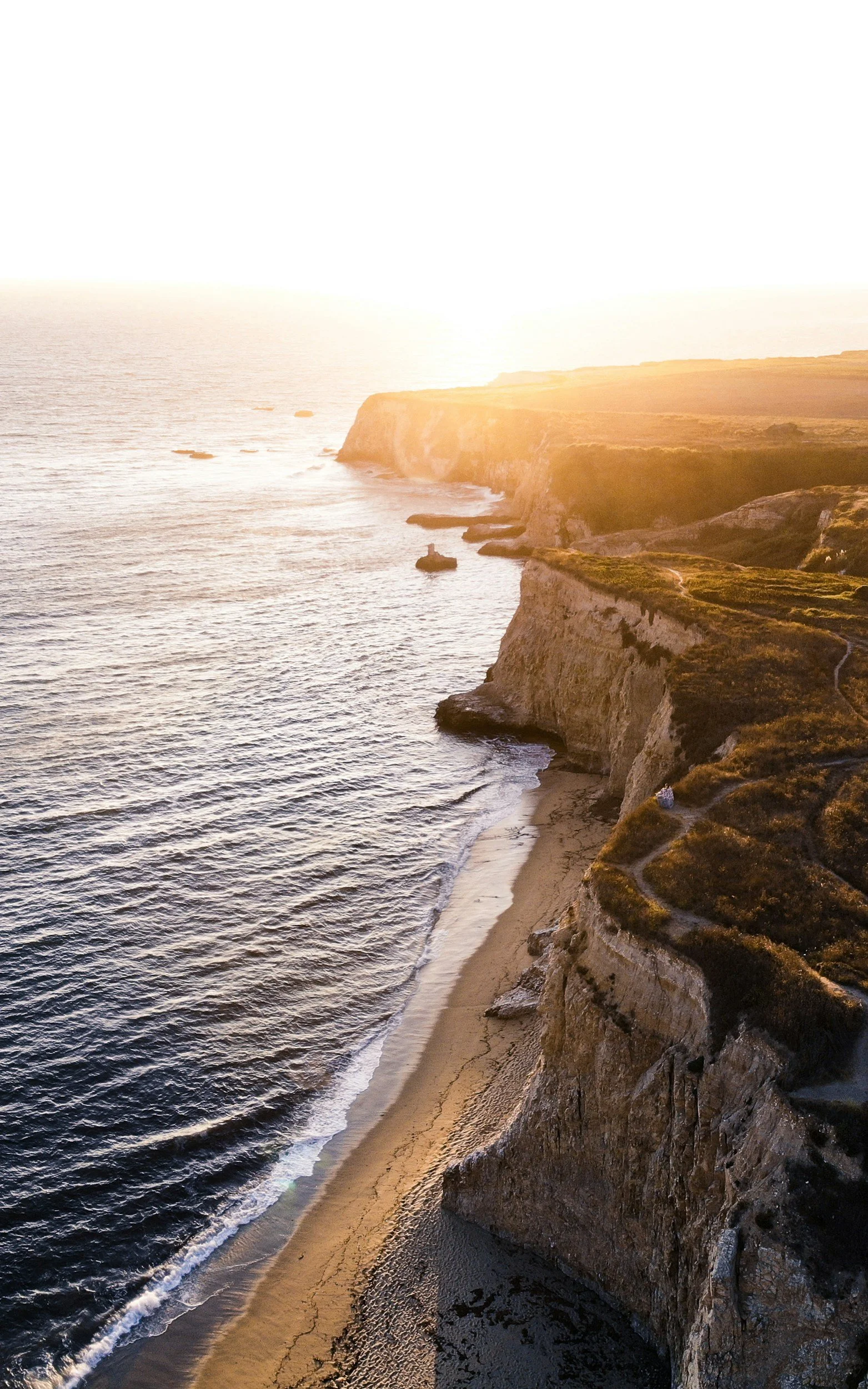 Sunset over rugged California coastal cliffs with a sandy beach below and ocean waves.