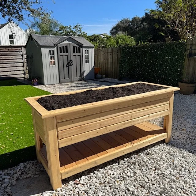 Custom cedar raised planter box with lower storage shelf, filled with organic soil and installed in a residential backyard garden.”