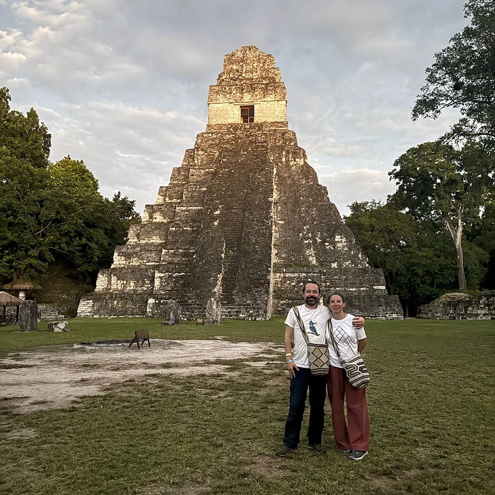 A man and woman standing together in front of a large ancient stepped pyramid at sunset, surrounded by trees, with clouds in the sky.