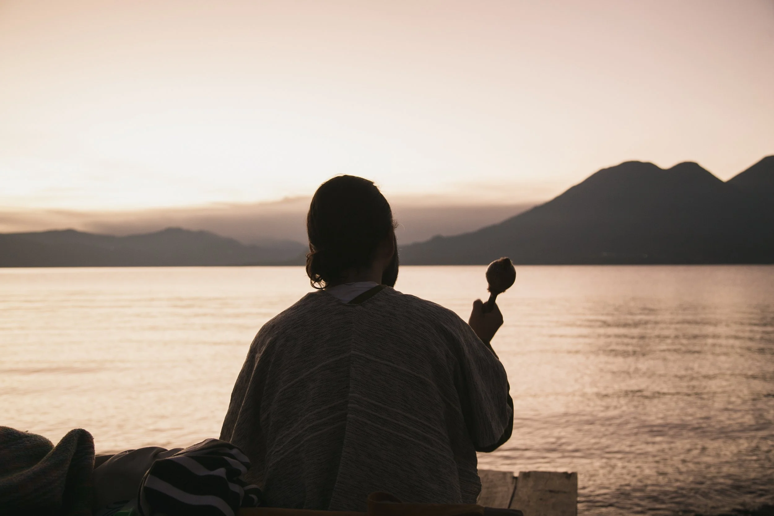 A person sitting by a lake at sunset, holding an ice cream cone with mountains in the background.