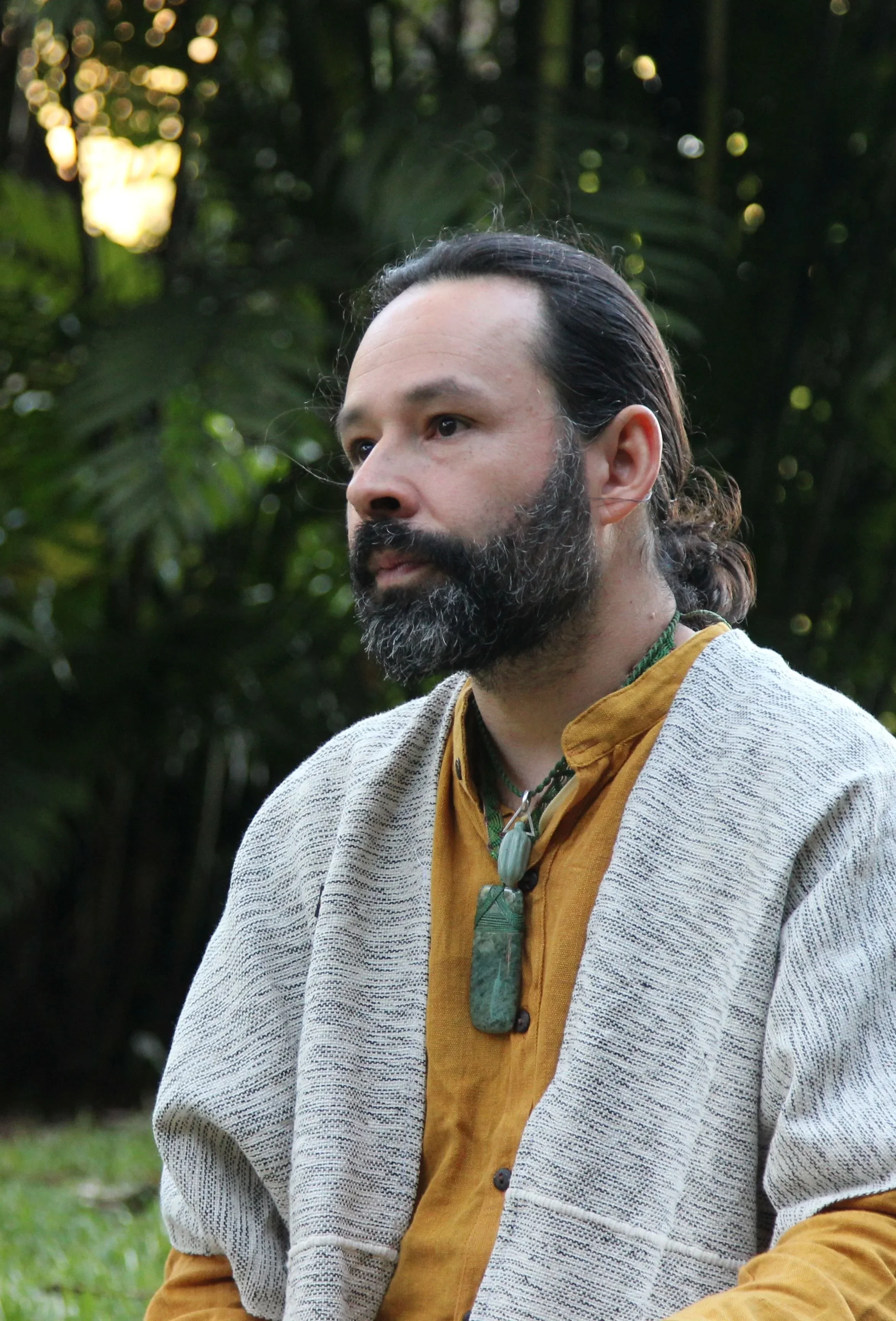 A bearded man with long dark hair, wearing a mustard yellow shirt and a light gray sweater, stands outdoors in a forested area with greenery and sunlight in the background.