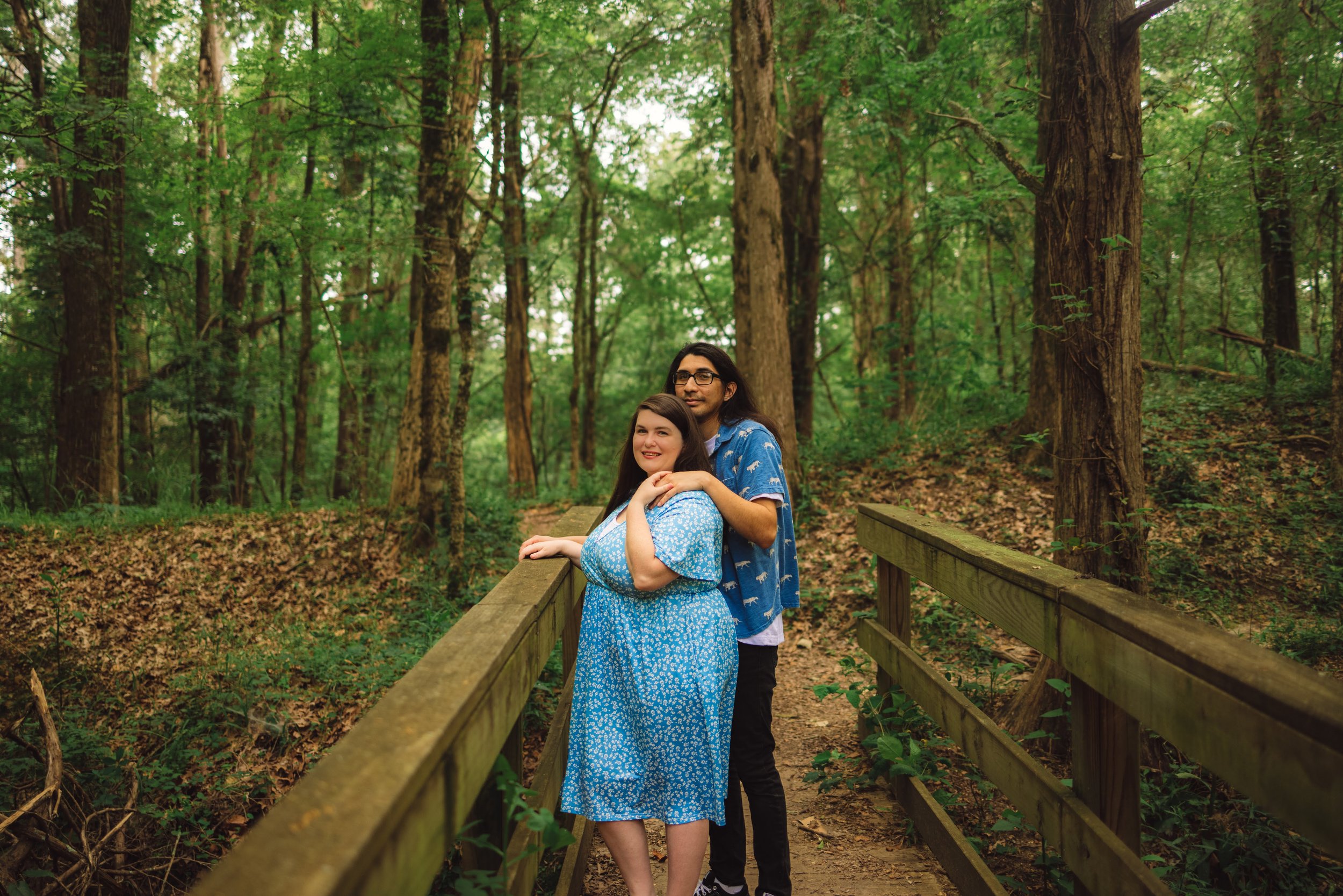 A couple standing on a wooden bridge in a lush green forest. The woman is wearing a blue dress and leaning on the bridge railing, while the man stands behind her with his hands on her shoulders. Trees and greenery surround them.