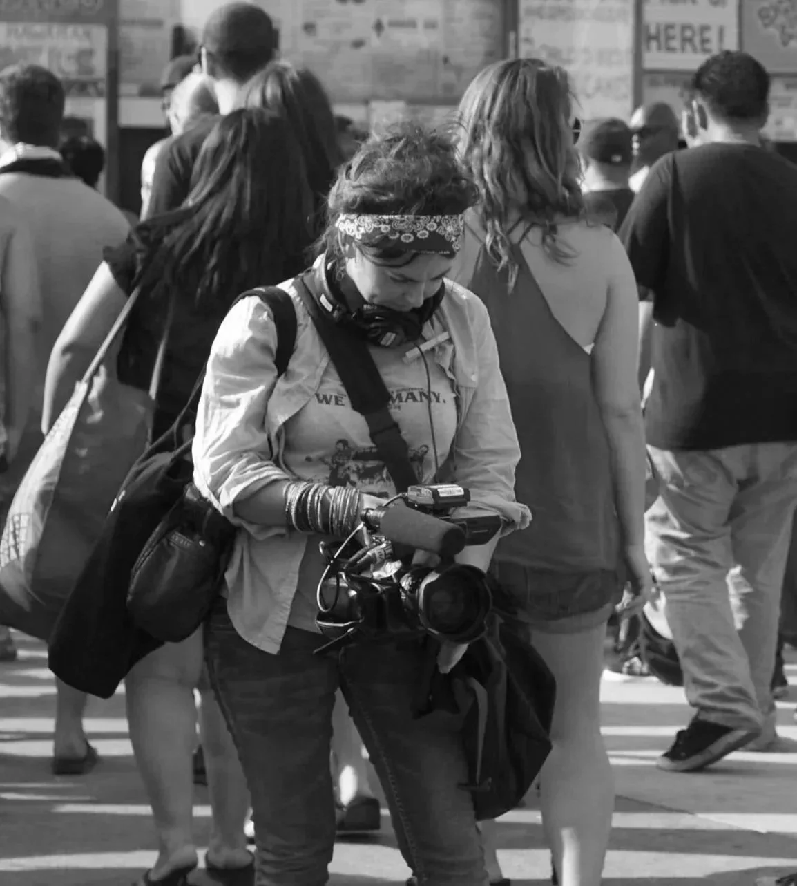 A woman with a camera and headphones around her neck walks through a crowd on a city street, looking at her phone.