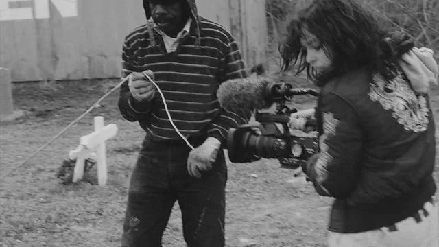 Two people and a dog in an outdoor yard, with one person holding the dog and the other person appearing to adjust camera equipment.