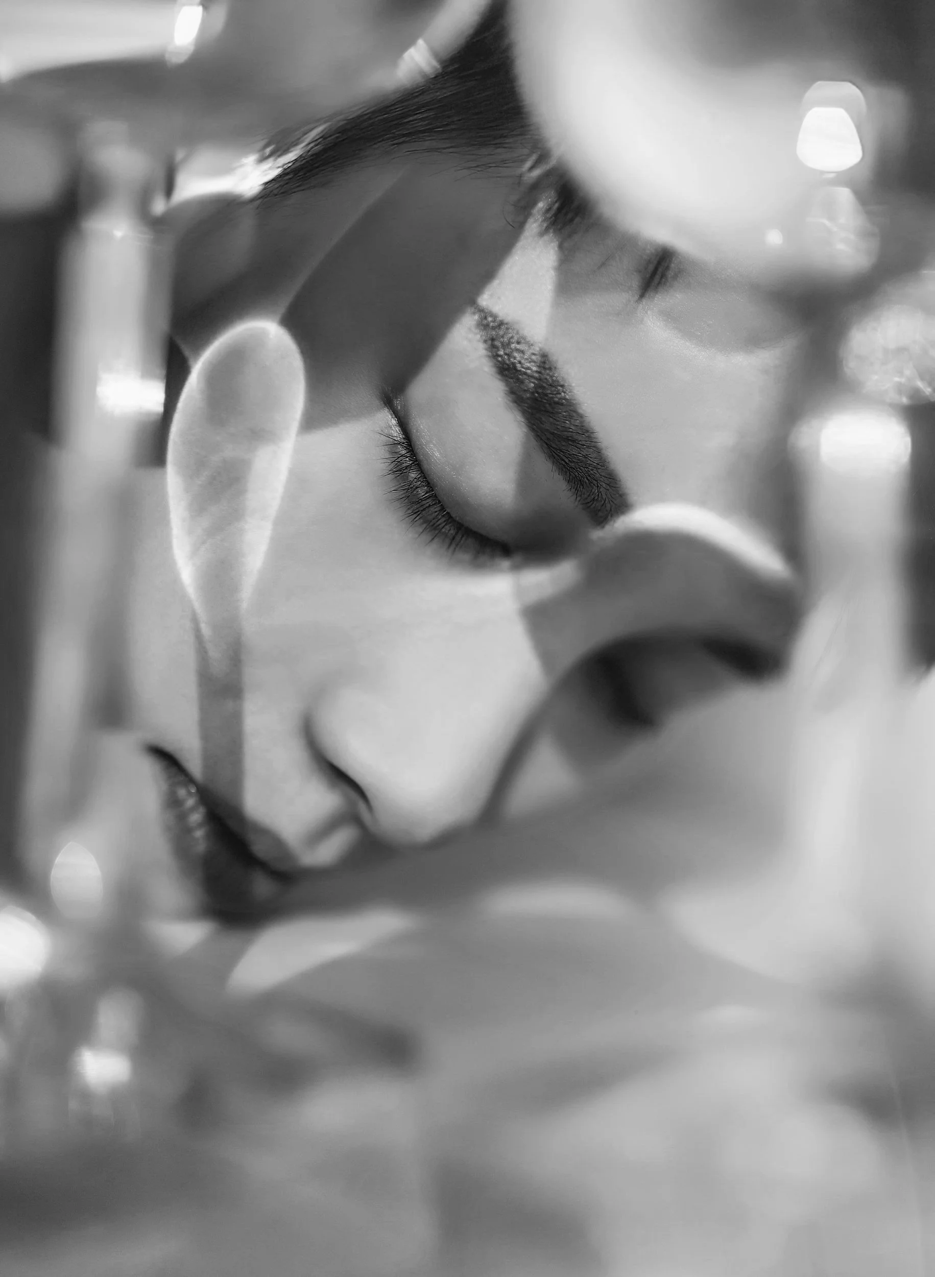 Close-up black and white photo of a person's face with closed eyes, viewed through glasses with reflections.