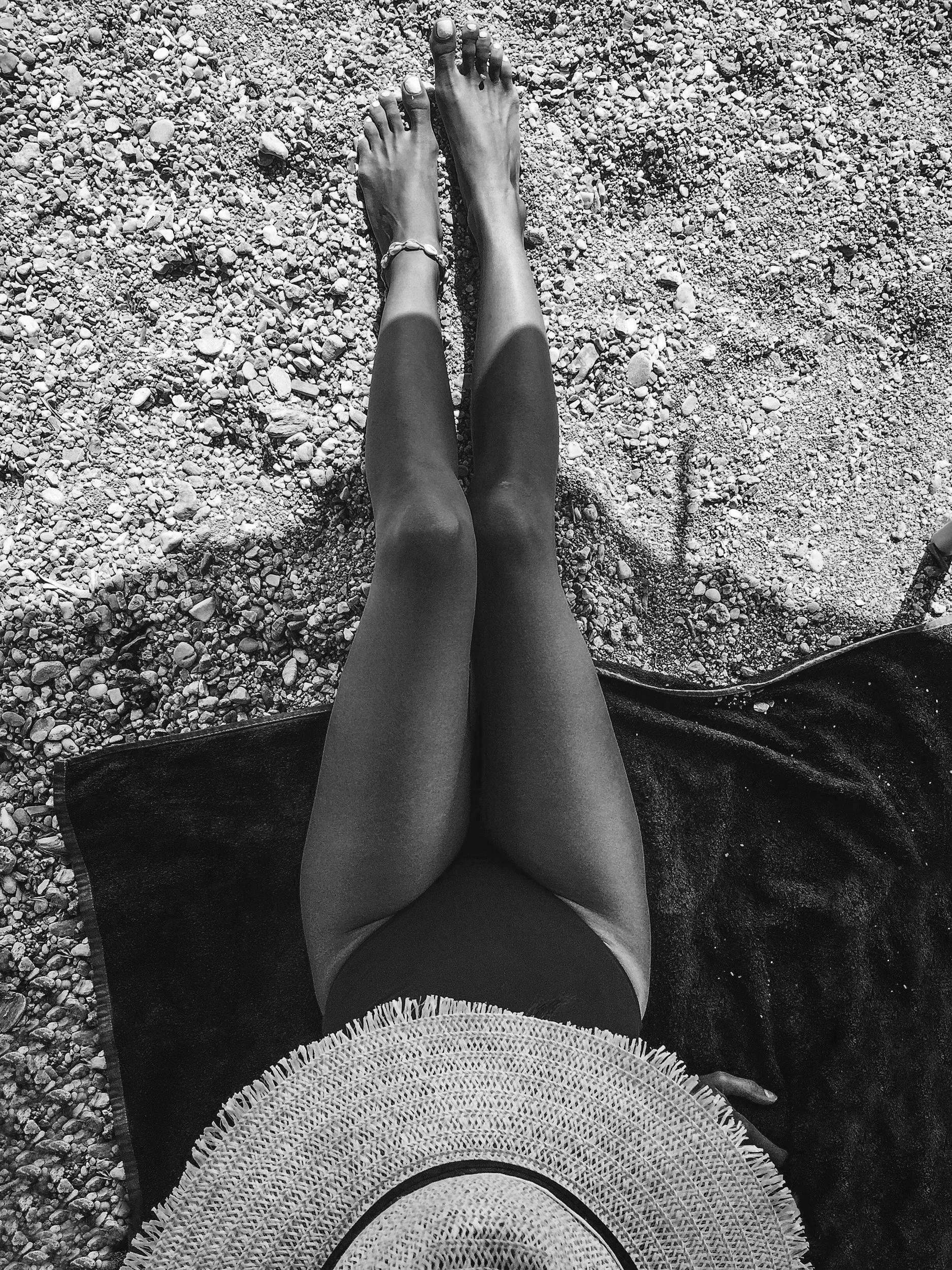 A person lying on a towel on a rocky beach, wearing a wide-brimmed straw hat, black swimsuit, and a bracelet, with their legs extended on the pebbled ground.