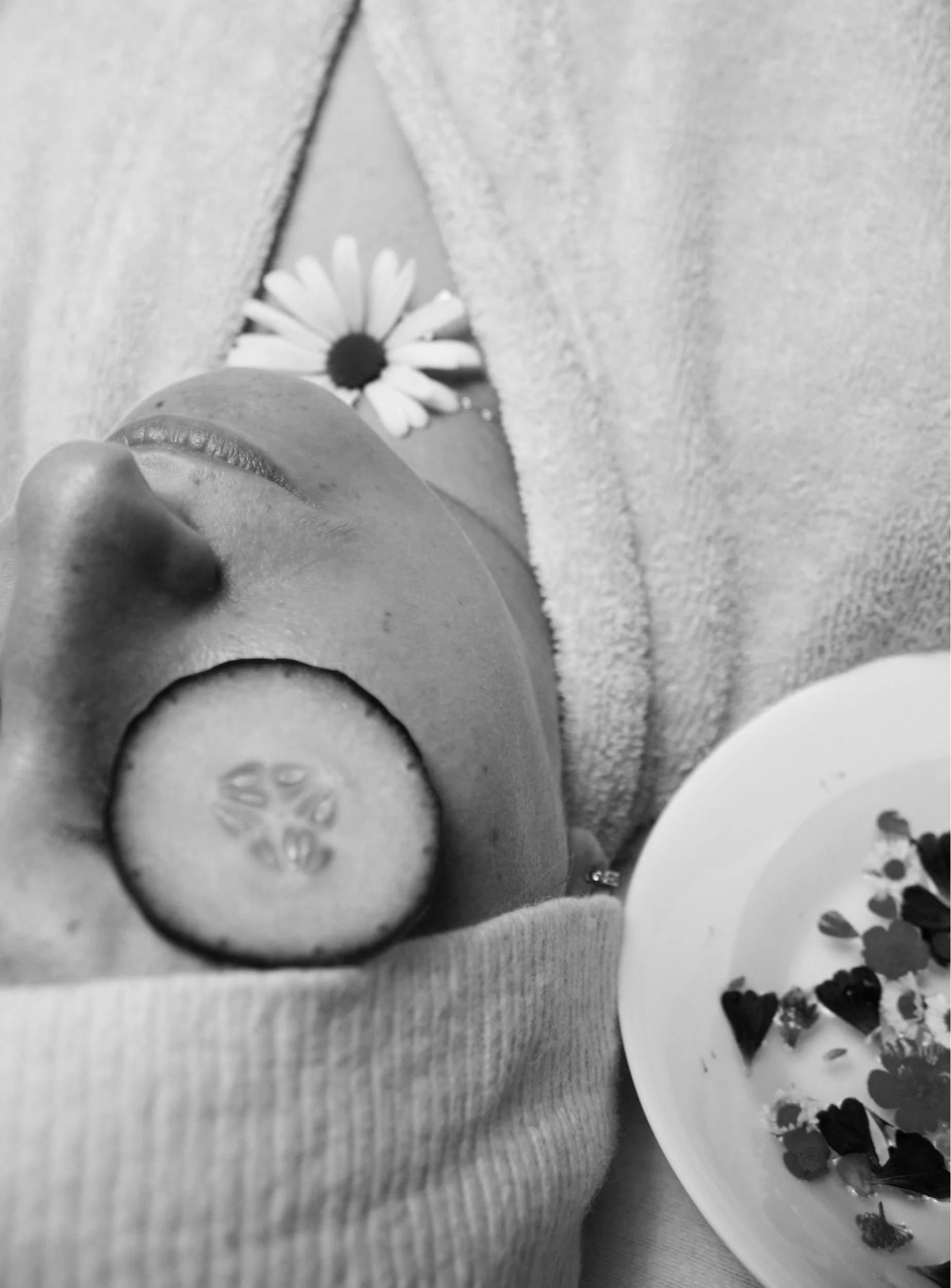 A close-up black and white photo of a person's face with a daisy flower behind their ear. They are holding a round slice of cucumber with seeds visible, near their lips. Part of a bowl with small, dark, heart-shaped decorations is visible in the lower right corner.