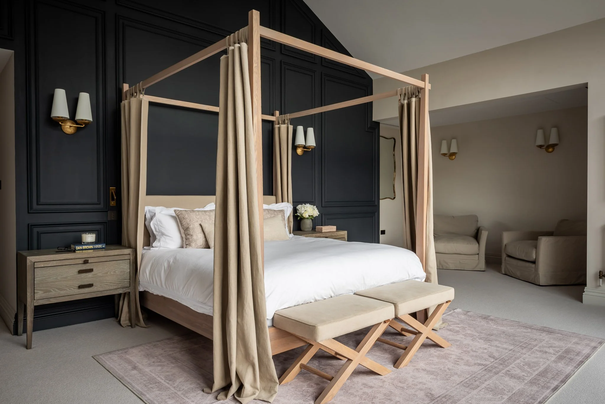 A luxurious bedroom with a four-poster bed, beige curtains, dark accent wall, two nightstands, and a sitting area with armchairs.
