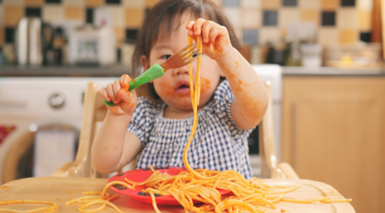 Toddler putting spaghetti on her fork with tomato sauce on her face