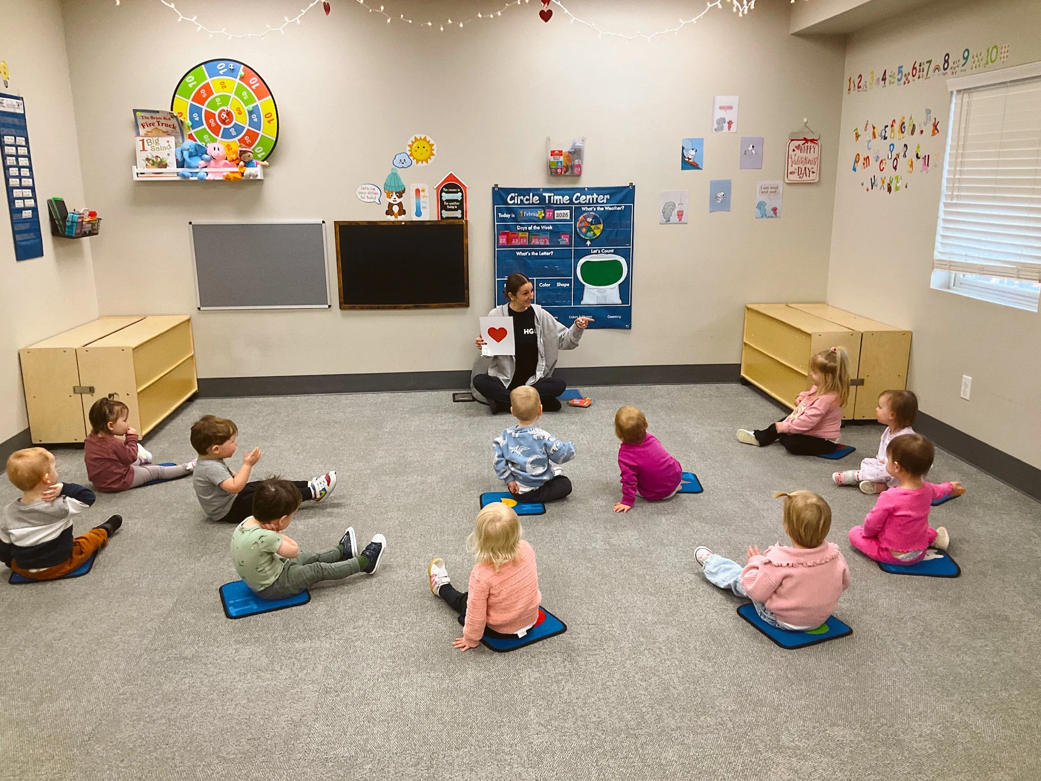 A woman leading a circle time activity for young children sitting on the floor in a classroom. The woman is holding a heart-shaped sign, seated cross-legged, surrounded by children sitting on individual mats or tiles. The classroom has educational posters, toys, and colorful decorations including a large clock on the wall.