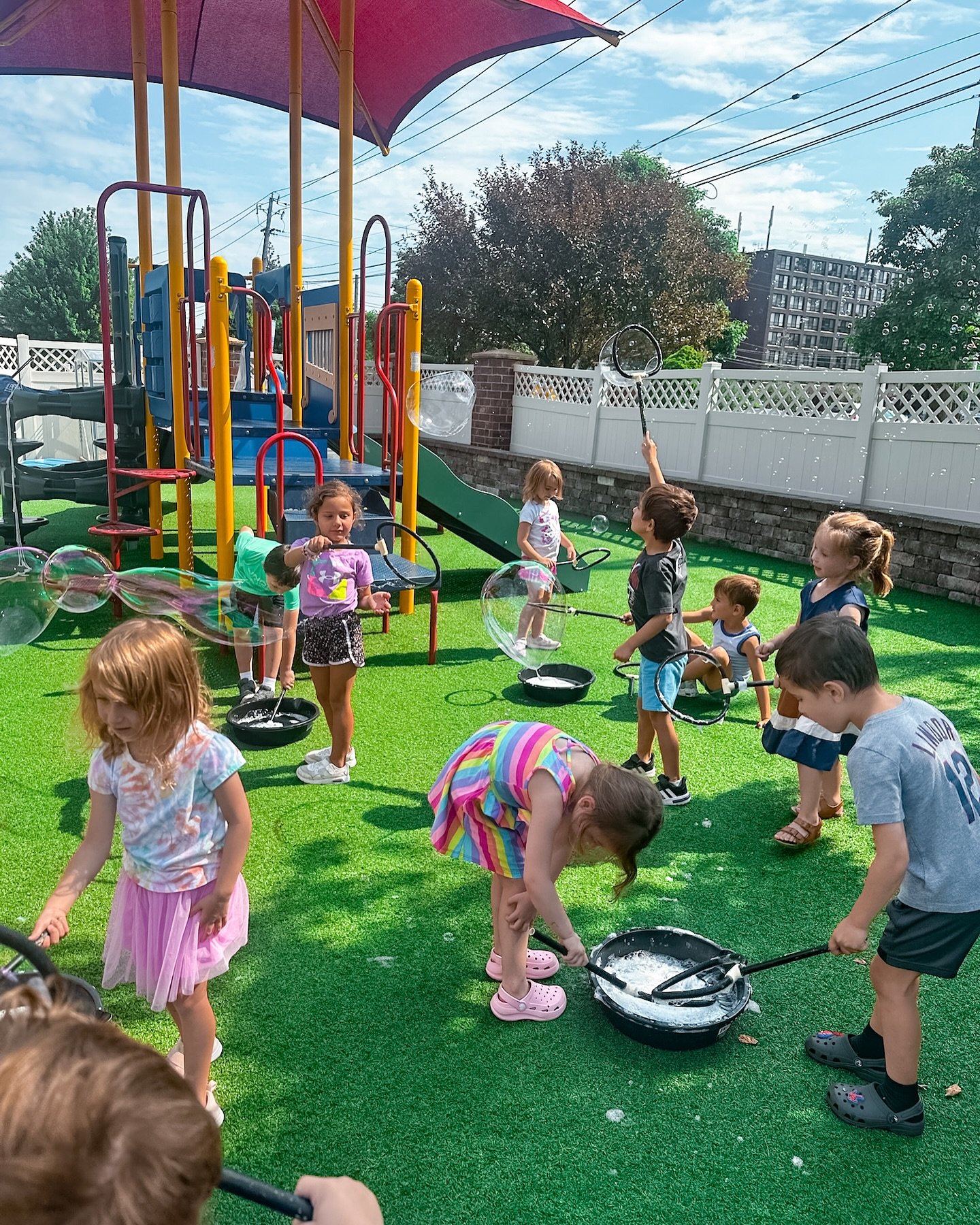 Kids playing with bubbles in a backyard play area with a slide, bubble bowls, and a white fence under a blue sky.