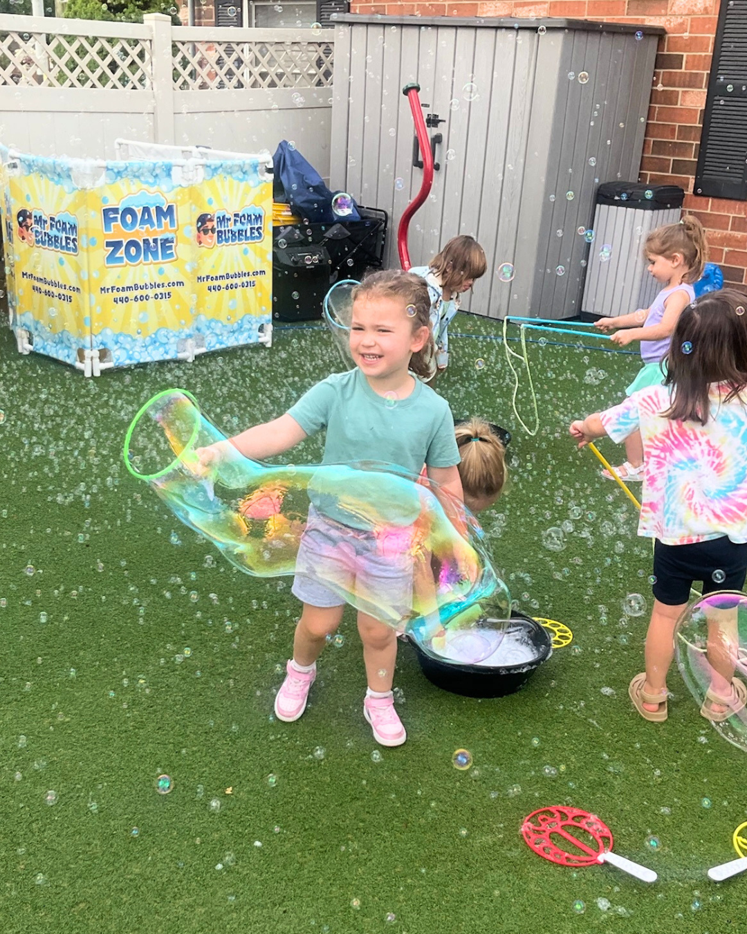 Group of children playing with bubbles in a backyard, with a foam bubble machine, slide, and fence in the background.