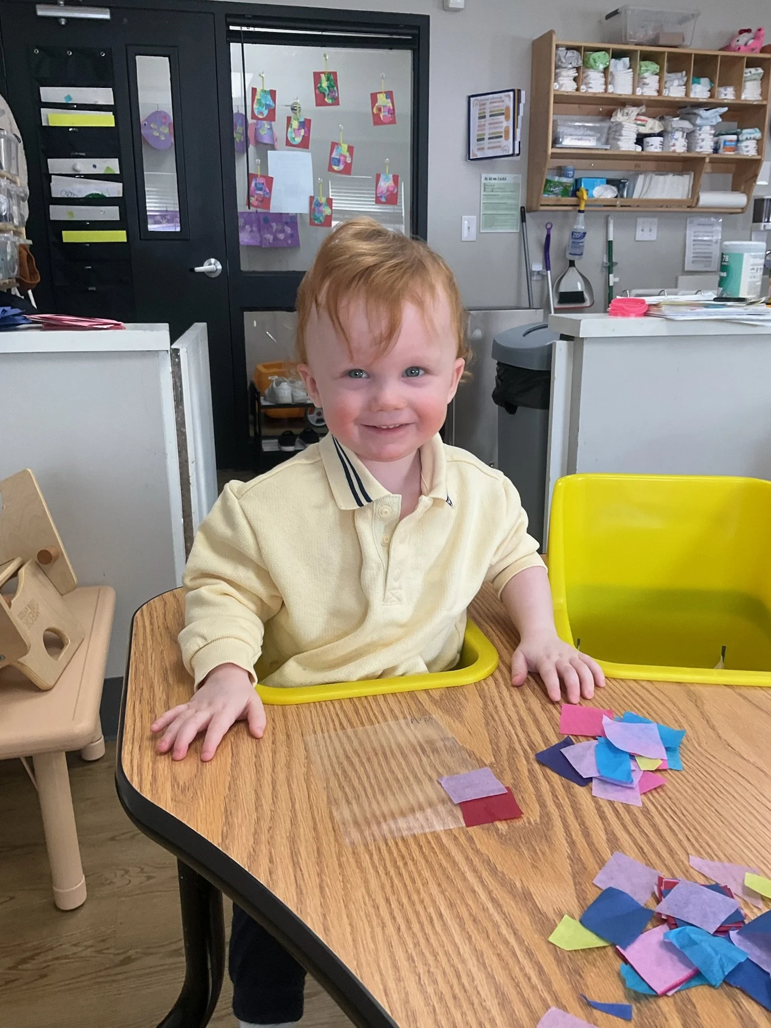 A young boy with red hair and light skin smiling at the camera, sitting at a wooden table lined with colorful pieces of tissue paper. He is wearing a yellow shirt and is in a room that appears to be a classroom or art room, with shelves of supplies in the background.