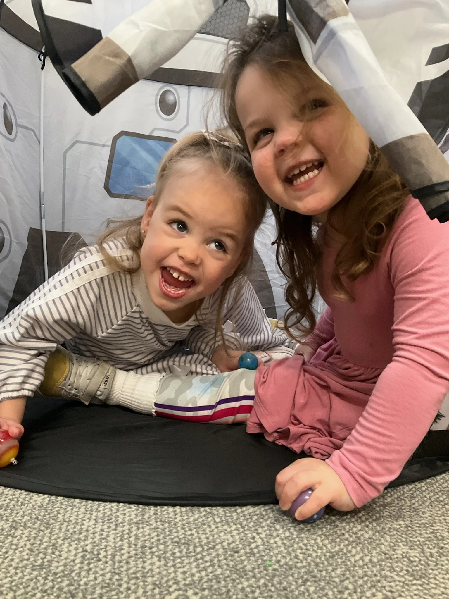 Two young girls sitting on the floor, smiling and enjoying a moment together. One girl has curly hair and is wearing a pink long sleeve shirt, the other girl has straight hair and is wearing a striped shirt. They are inside a tent.