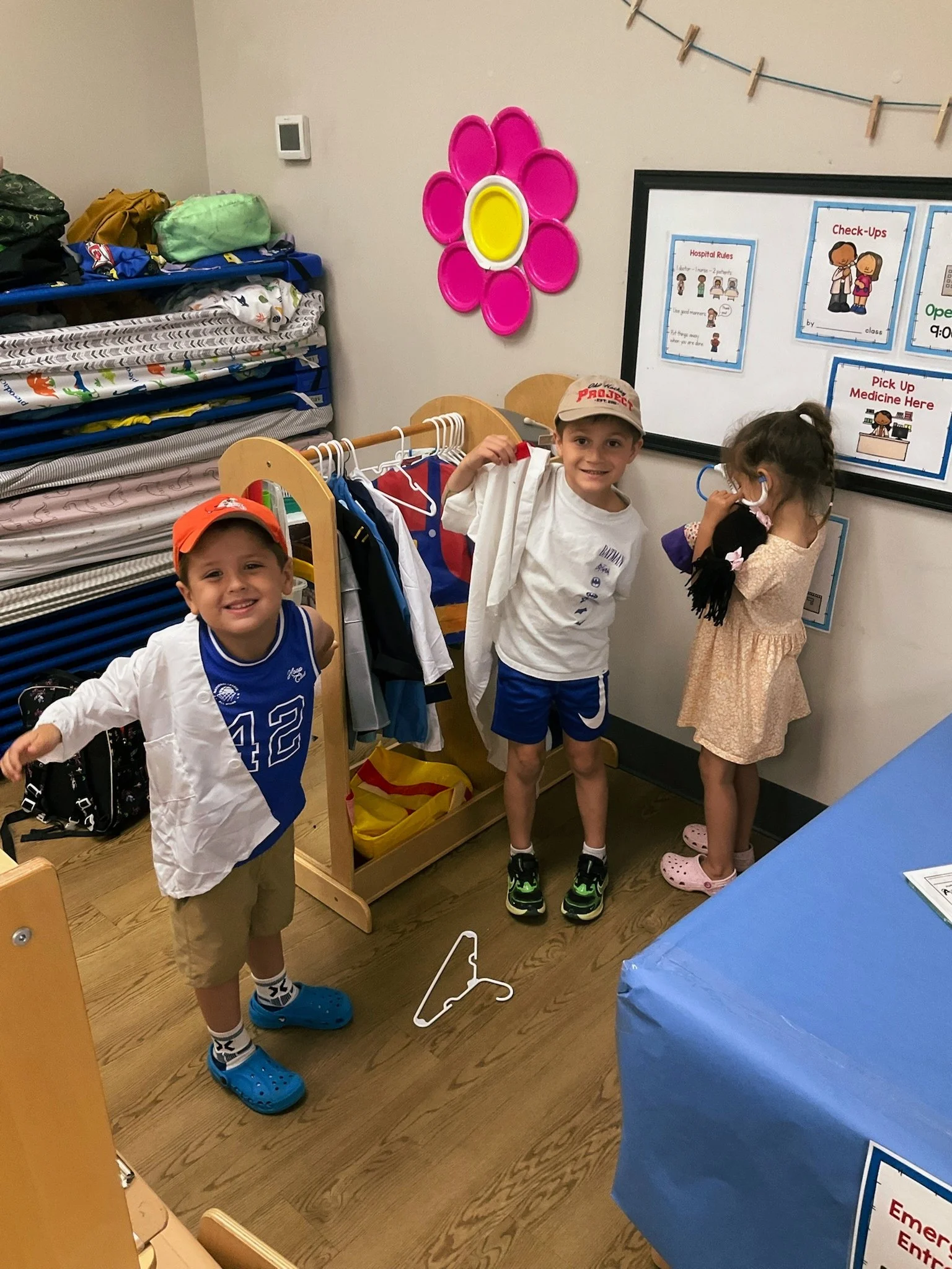 Three children standing in a room near a coat rack, some clothes hanging, and a whiteboard with hospital rules. One boy is smiling at the camera wearing a sports jersey and cap, another boy is holding a shirt, and a girl is listening to a stethoscope.