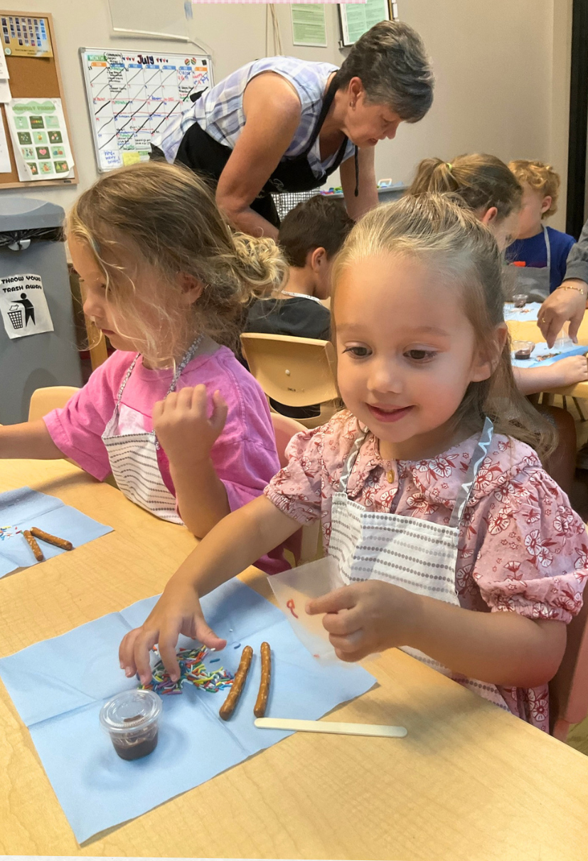 A young girl in a pink floral dress and striped apron is sitting at a table with blue napkin, decorating pretzels with colorful sprinkles and chocolate syrup. Other children and an adult are working on similar activities at the same table in a classroom setting.