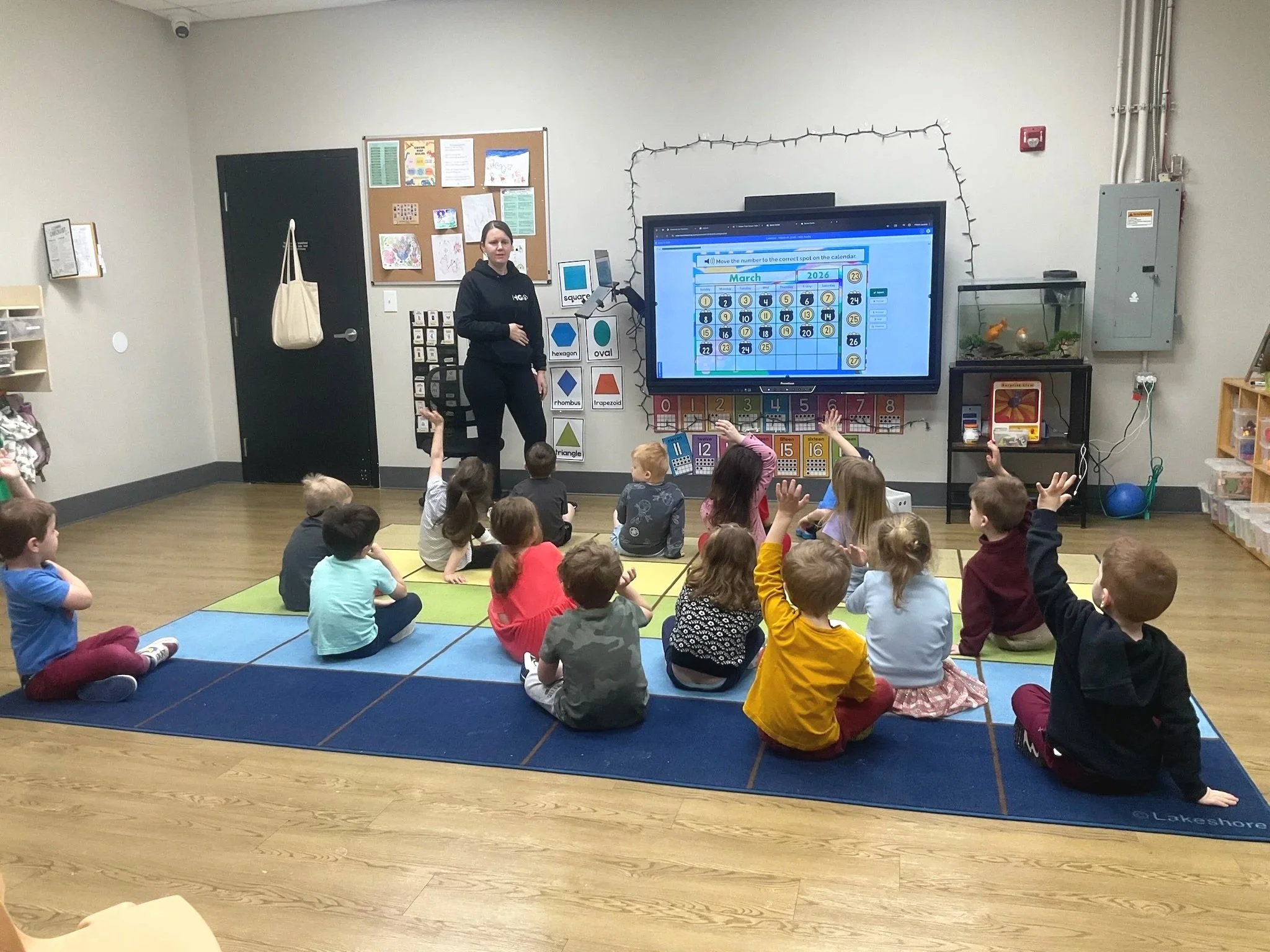 A classroom teaching session with young children sitting on a colorful mat, facing a teacher standing beside a large screen displaying a calendar activity. Some children have their hands raised.