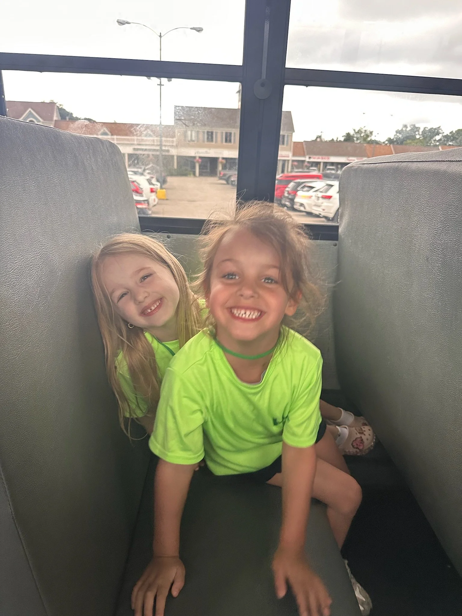 Two young girls with blonde and brown hair smiling, posing inside a transportation vehicle, with parked cars and buildings visible outside the window.