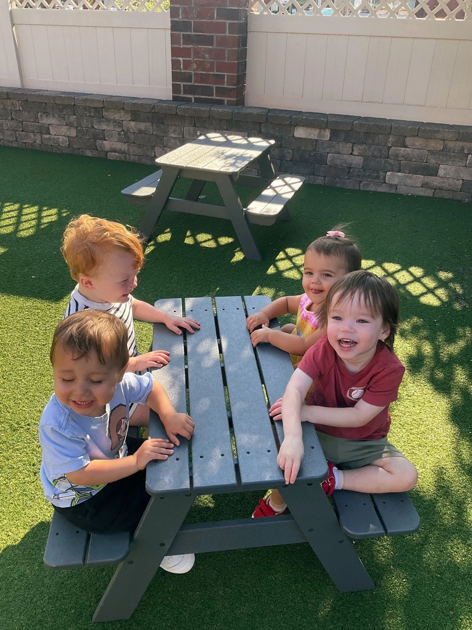 Six young children sitting around a gray wooden picnic table outdoors, with a brick and white fence in the background on a sunny day.