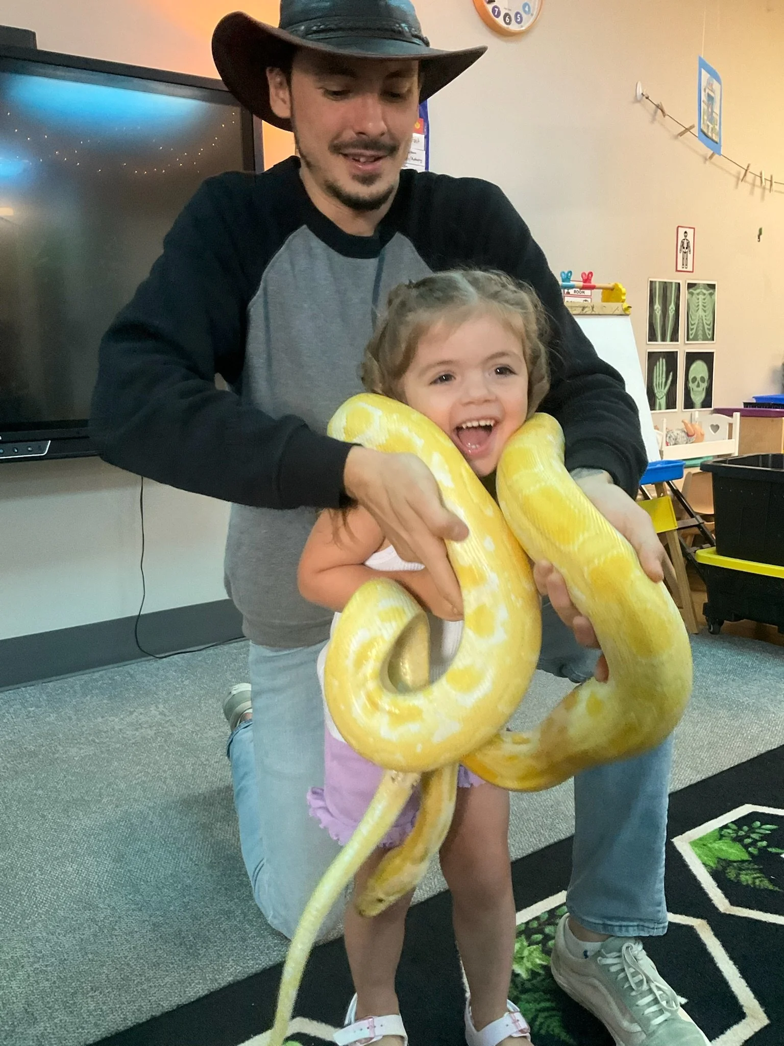 A man wearing a cowboy hat is holding a smiling young girl wrapped in a large yellow snake or boa constrictor inside a room with educational posters and classroom furniture.
