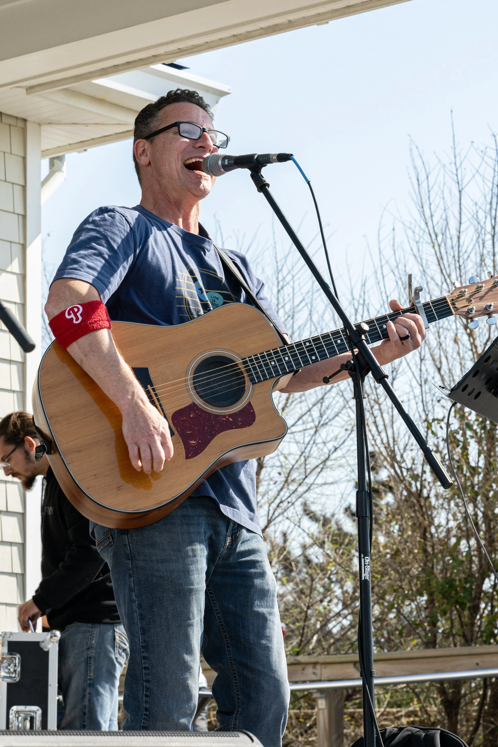 Man singing and playing guitar outdoors with a microphone, wearing glasses and a red arm band, during daytime.