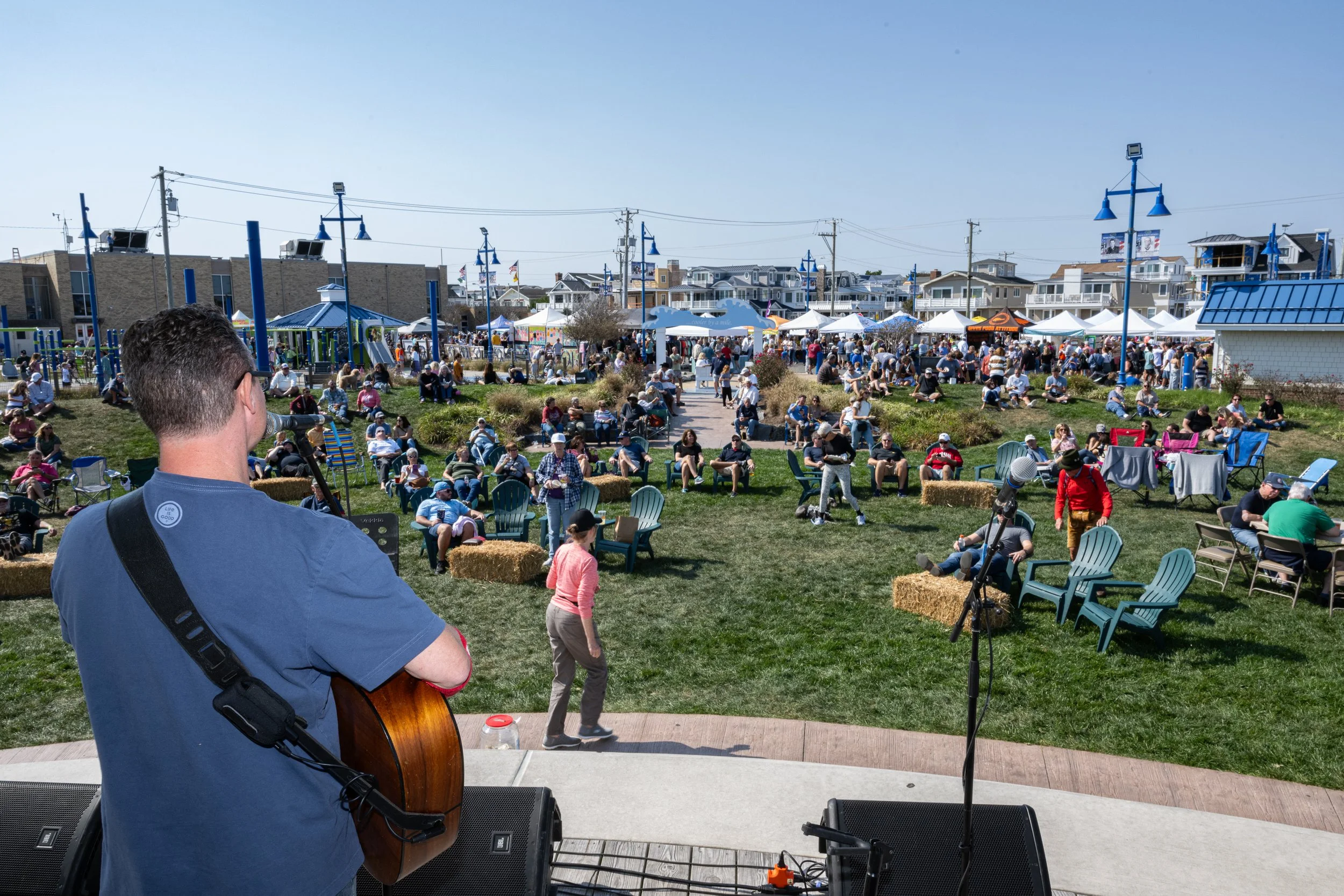 A musician playing guitar on stage at an outdoor event. The audience is seated on chairs and hay bales on a grassy area, with many people standing and socializing. Tents and houses are visible in the background on a sunny day.
