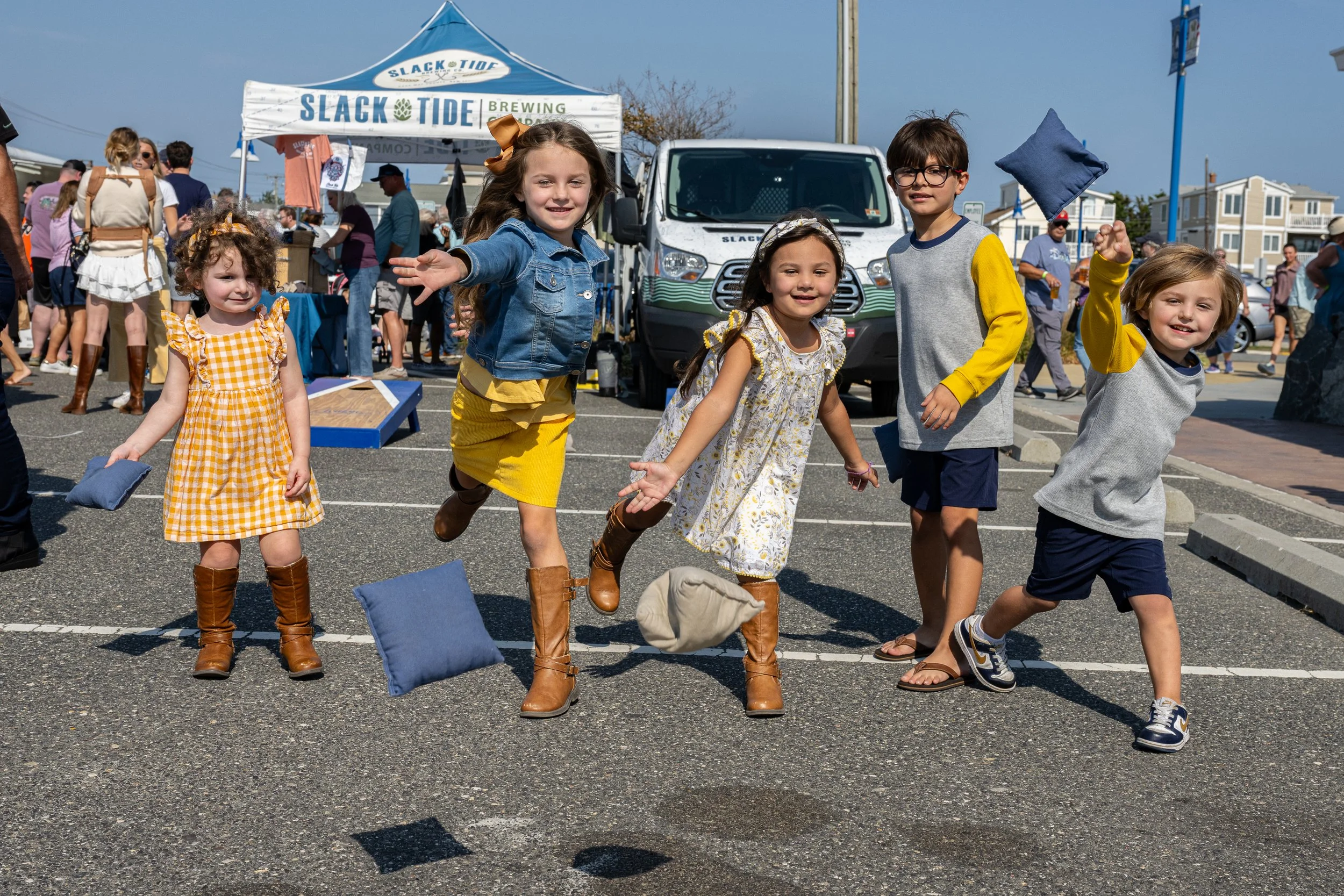 Several children playing a bean bag toss game at an outdoor event, with some children in mid-action and others holding bean bags, under tents and around a parking lot.