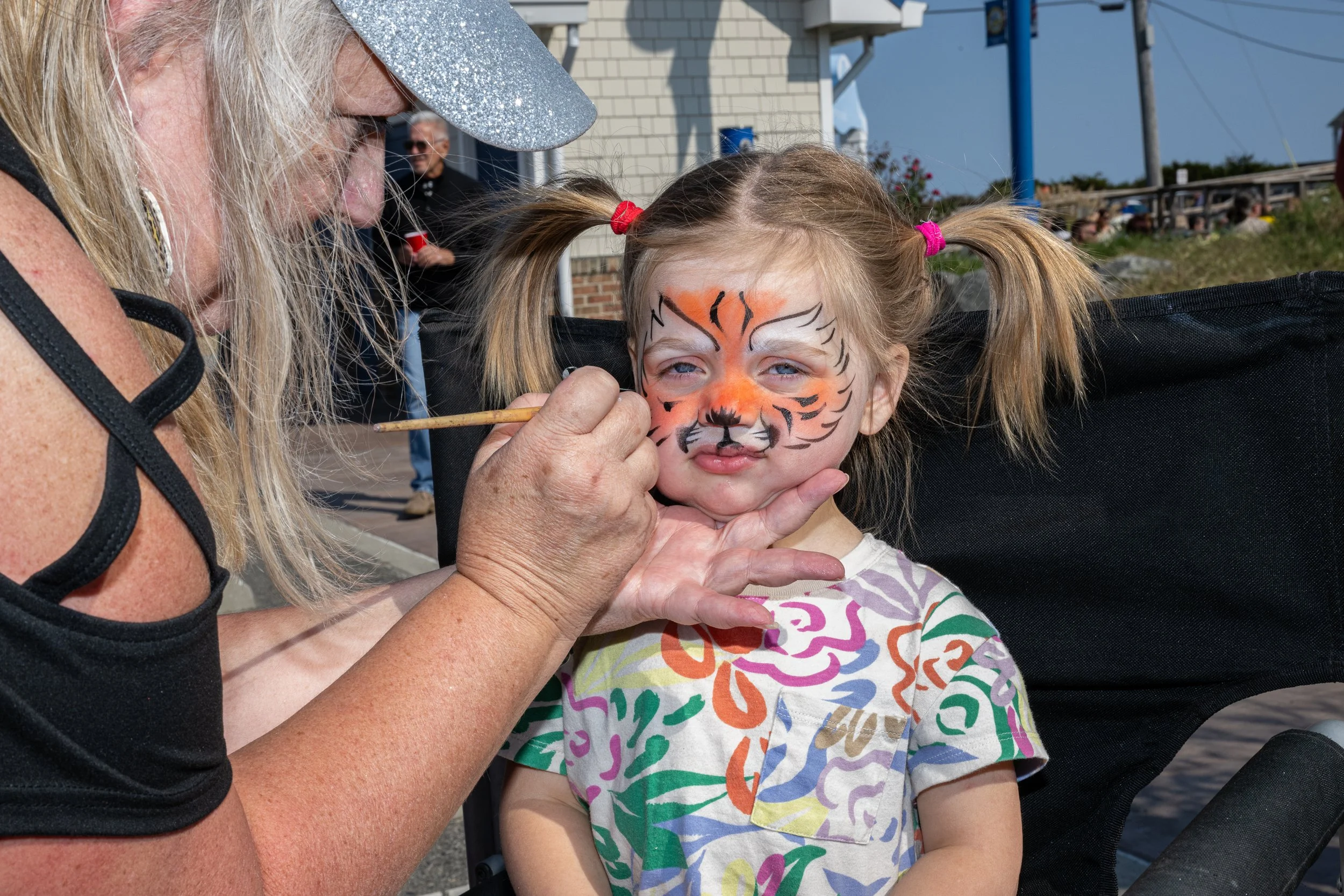 An older woman is painting a tiger face on a young girl's face during an outdoor event, with other people visible in the background.