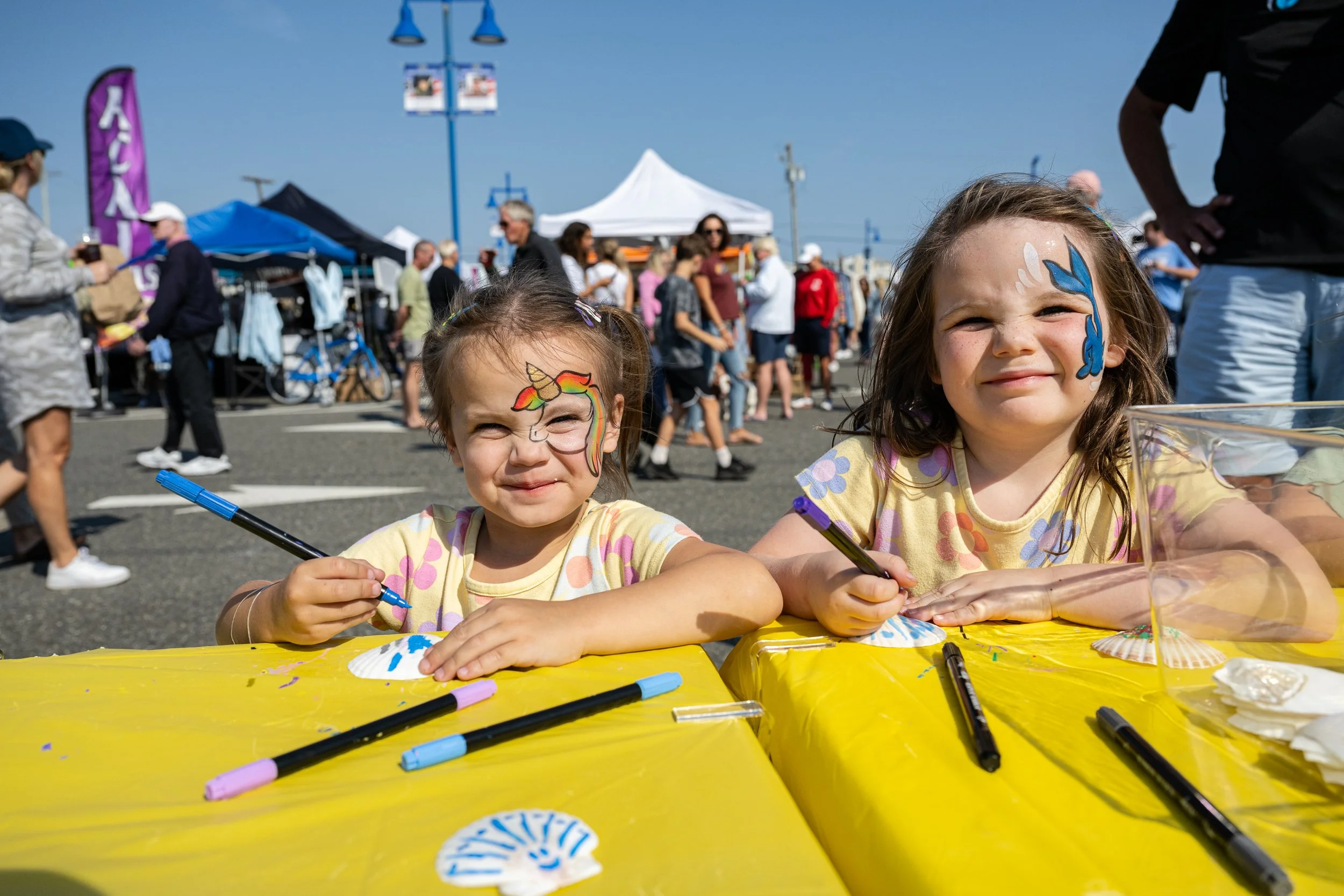 Two young girls with face paint of unicorn and blue horse, sitting at a yellow table, drawing with markers at an outdoor event with tents and people in the background.