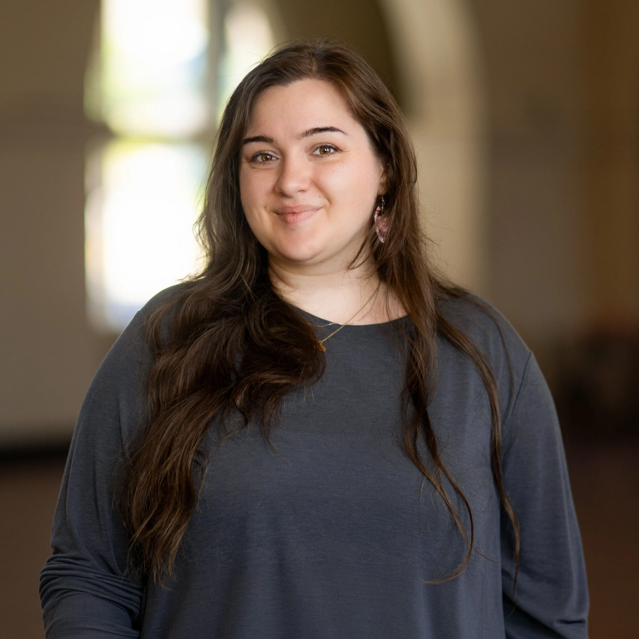 A young woman with long brown hair, fair skin, and a subtle smile wearing a dark blue shirt and pink beaded earrings.