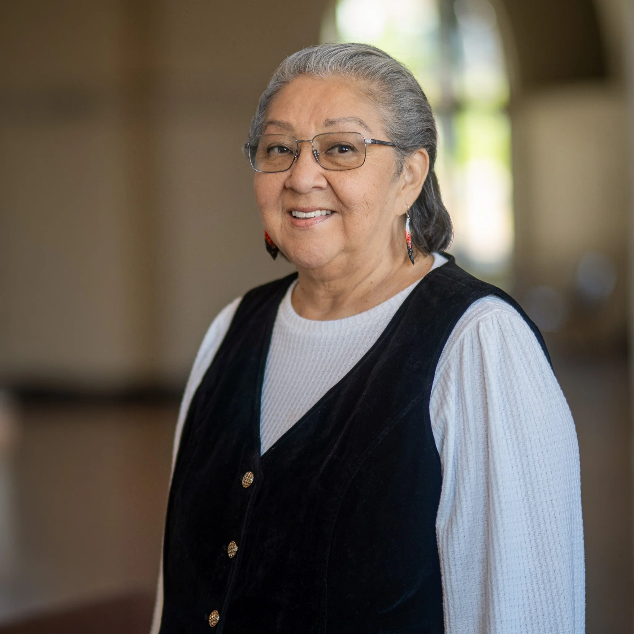 Woman with short gray hair wearing a white long sleeve shirt under a black velvet vest. She is also wearing beaded earings and eye glasses