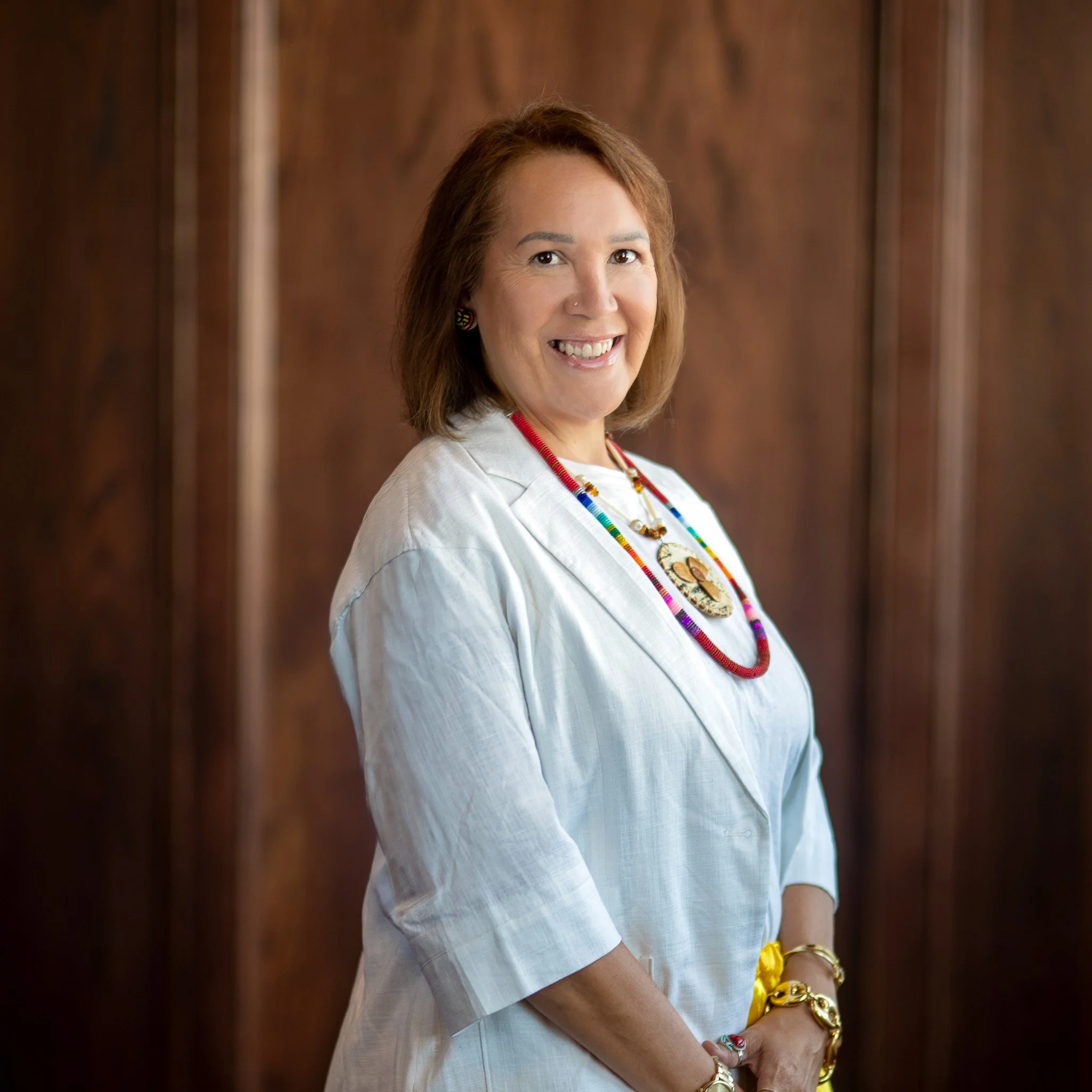 Portrait of a smiling woman with short brown hair, wearing colorful jewelry and a white shirt, against a wood background.