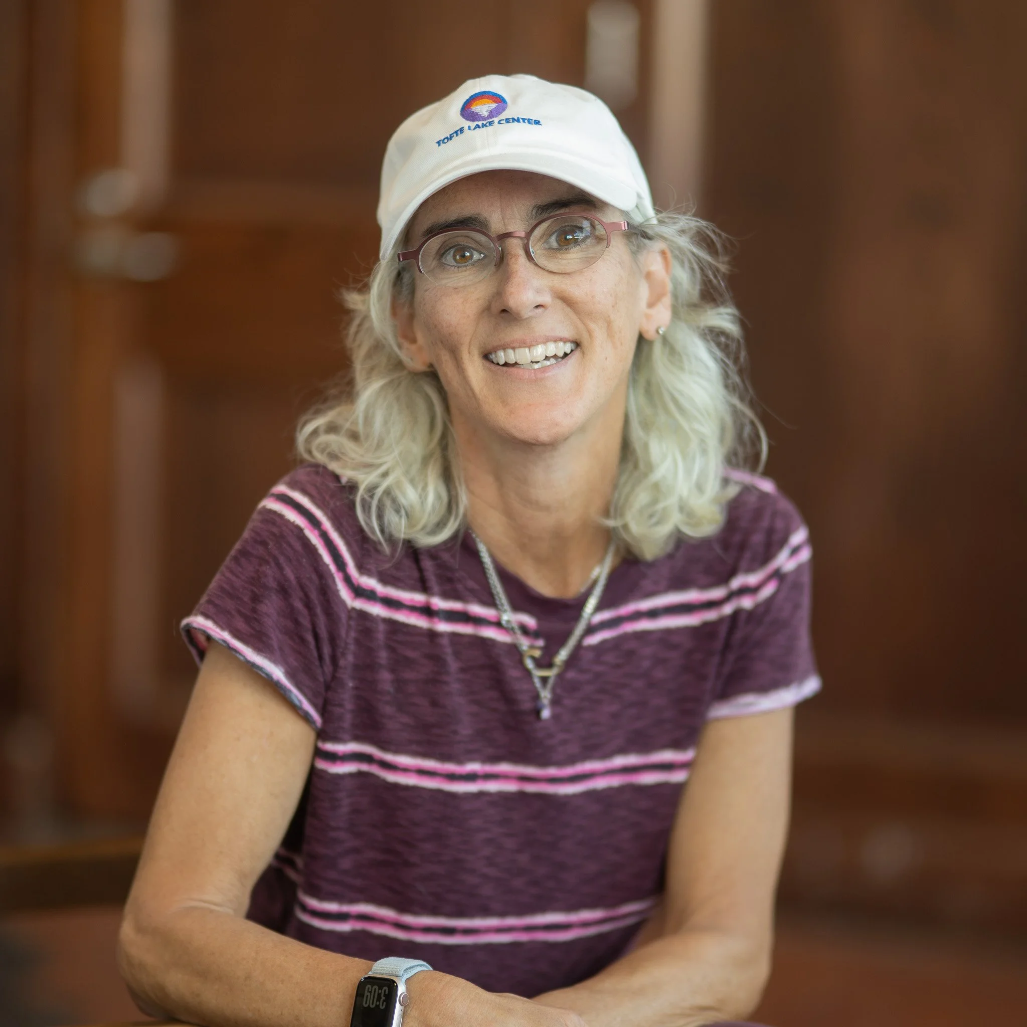 Smiling woman with long gray hair, glasses, and layered necklaces, wearing a purple patterned top with pink sleeves, standing in front of a colorful yellow and orange abstract background.