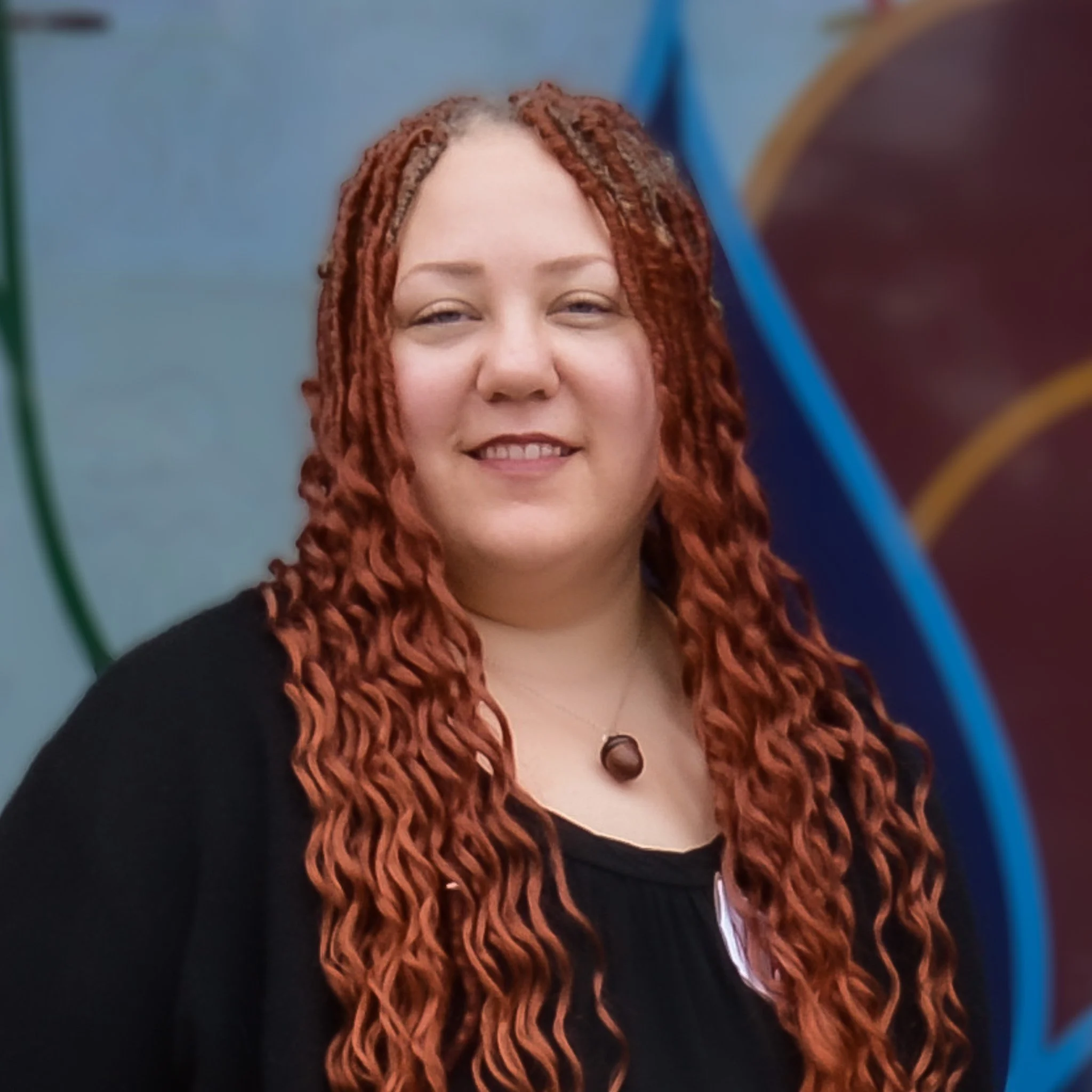 Portrait of a woman with long red curly hair and a black top, smiling, standing in front of an abstract yellow and orange background.