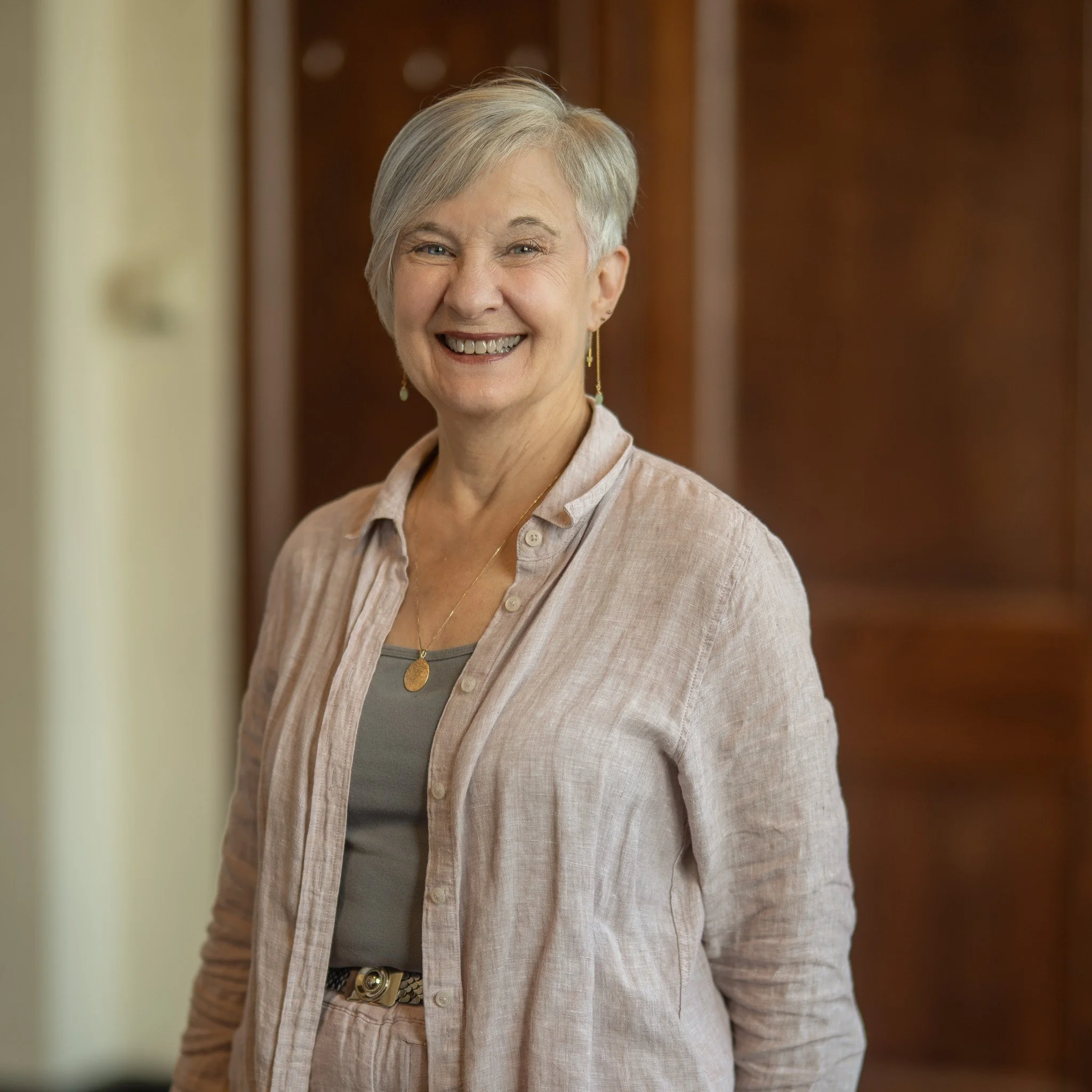 A smiling woman with short gray hair standing against a colorful abstract background, wearing a blue top and a light beige jacket.