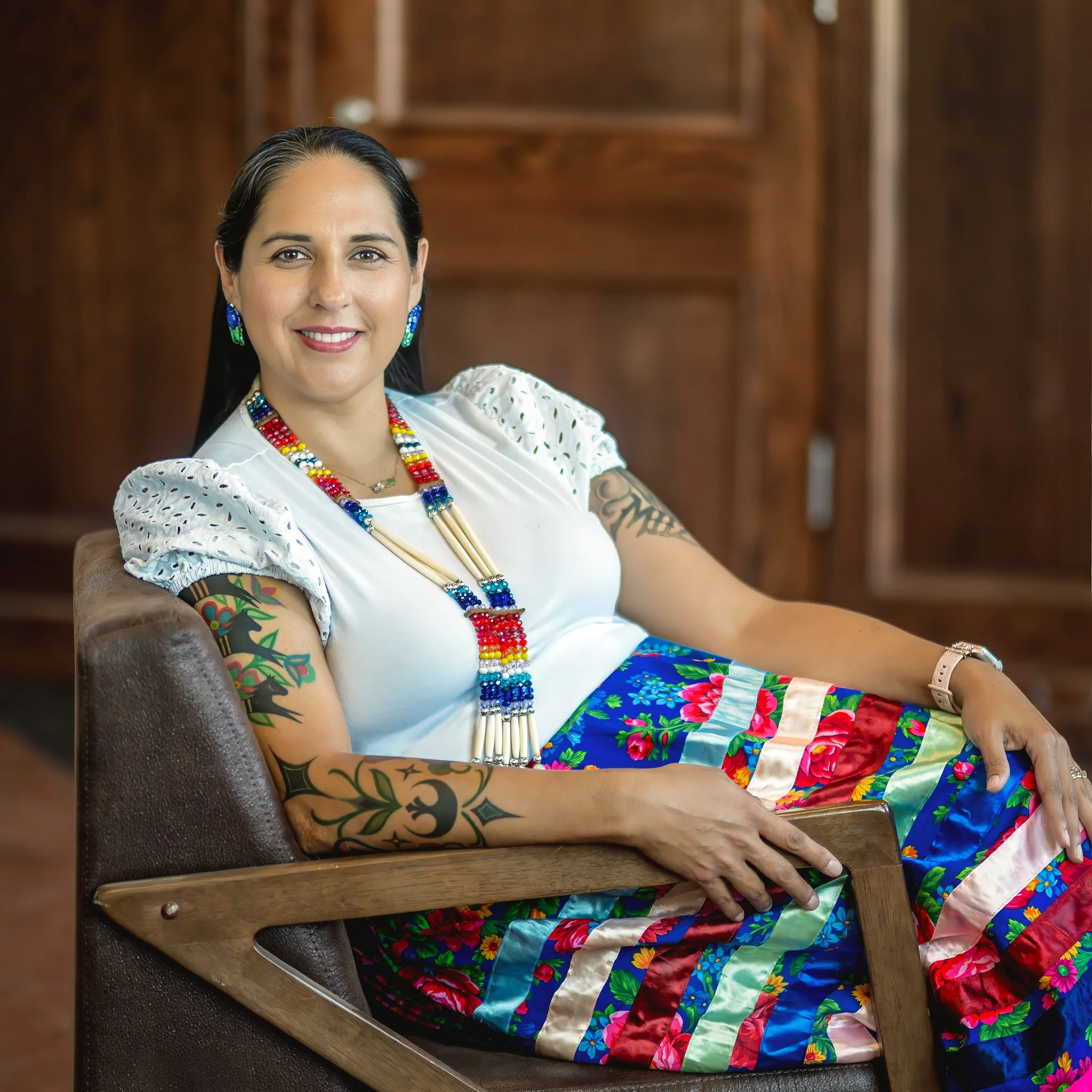 A woman smiling, wearing a white shirt, blue denim jacket, and colorful beaded jewelry, standing in front of a yellow artistic background.