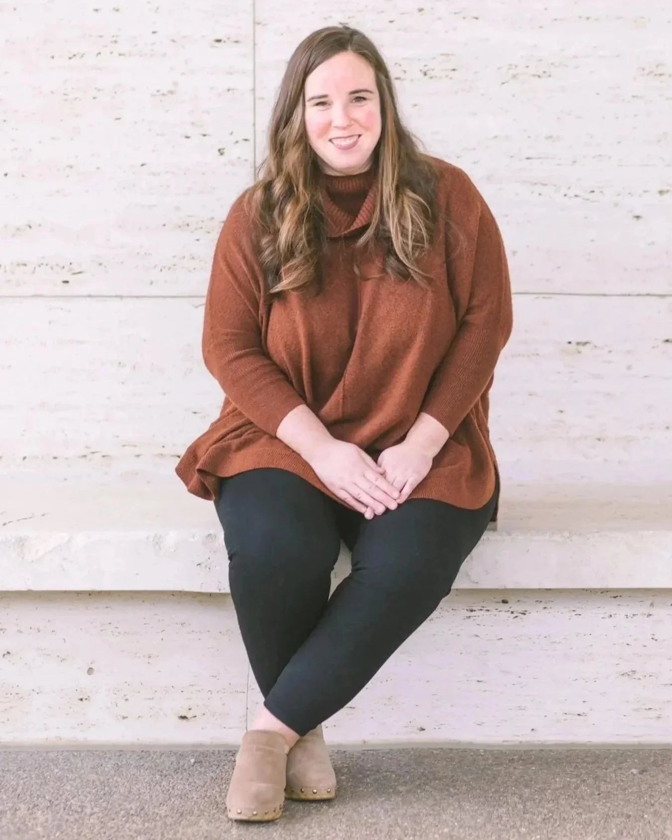 A woman with long, wavy brown hair sitting on a white marble bench in front of a white marble wall, wearing a rust-colored sweater, black pants, beige ankle boots, and smiling at the camera. Emily M. Lewis Counseling.