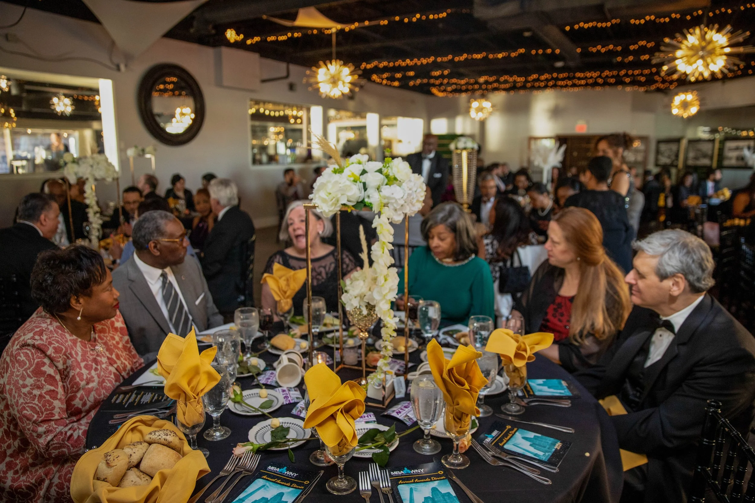 A group of people in formal attire gathered around a decorated dining table at an event with floral centerpieces, yellow napkins, and a festive ceiling with string lights and chandelier-style fixtures.