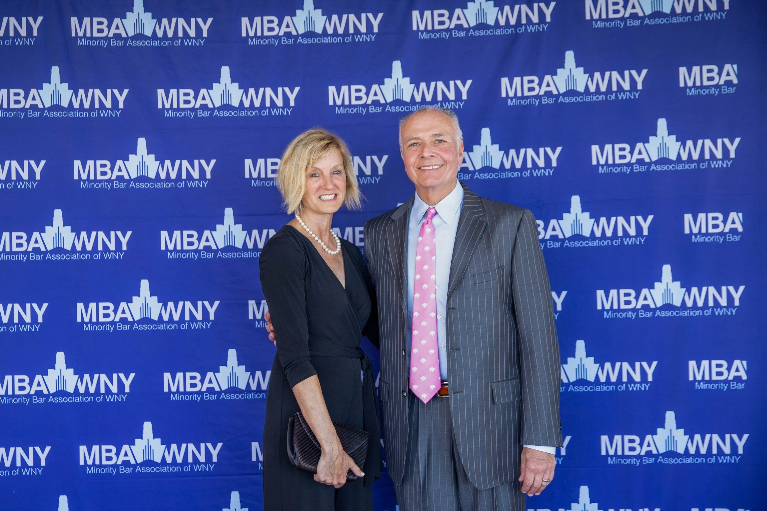 A man and a woman are standing together in front of a blue backdrop with the logo and name of the Minority Bar Association of WNY. The woman has blonde hair, is wearing a black dress and pearl necklace, and is holding a black clutch. The man has gray
