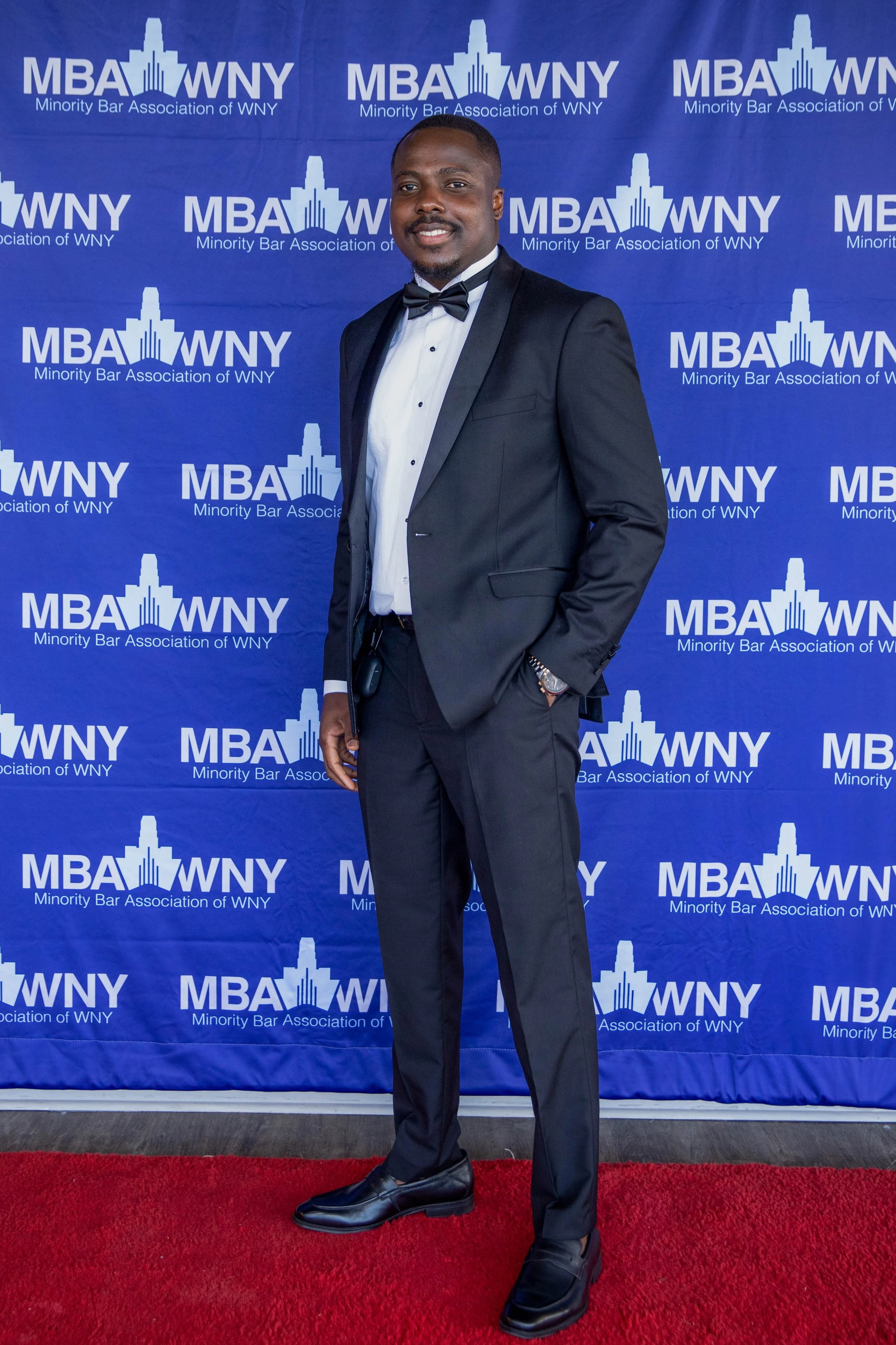 A man in a formal black tuxedo with a bow tie is standing on a red carpet in front of a blue backdrop. The backdrop has the logo and text for the Minority Bar Association of WNY, with a city skyline graphic and the initials "MBA WNY" repeated multipl